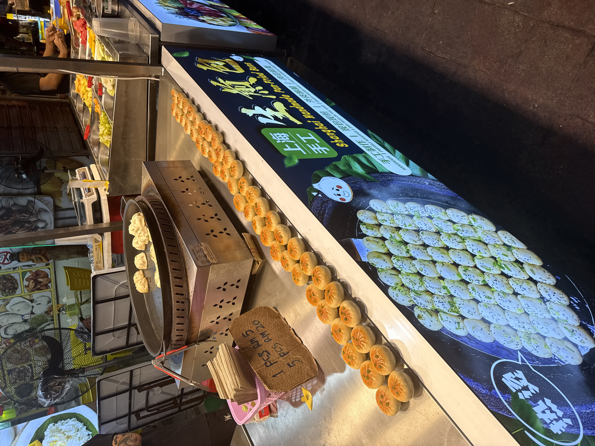 This nighttime image captures a vibrant street food stall, likely in a night market in Kuala Lumpur, Malaysia. The main focus is on the preparation and display of "Shanghai Handmade Pan-Fried Buns."
In the foreground, rows of golden-brown, pan-fried buns, often garnished with green specks, are neatly arranged on a metallic counter. To their left, a large, circular griddle is actively cooking more buns, suggesting fresh preparation. Adjacent to the griddle is a metal unit with decorative cutouts.
A prominent, illuminated sign stretches along the counter, displaying "Shanghai Handmade Pan-Fried Buns" in both English and Chinese characters ("上海手工"). The sign also includes descriptive text: "HANDMADE / FRESHLY WRAPPED & PAN FRIED ON THE SPOT" and "手工制作 / 现包现的外皮酥策、内运附室," which translates to handmade, freshly wrapped and pan-fried, with crispy skin and rich filling. Images of both the golden-brown buns and a flatter, sesame-and-scallion topped variety are featured on the sign.
A handwritten cardboard sign clearly indicates the pricing: "1 PCS RM 5" and "5 PCS RM 20," confirming the use of Malaysian Ringgit. A pink plastic basket holds stacks of paper packaging, ready for customers.
In the background, other food stalls are visible with various ingredients, such as colorful fruits or vegetables in metal trays. A person's hands are seen working at an adjacent stall. A large black industrial fan suggests a warm environment. The bright artificial lighting throughout the scene highlights the food and activity, creating an inviting night market atmosphere.