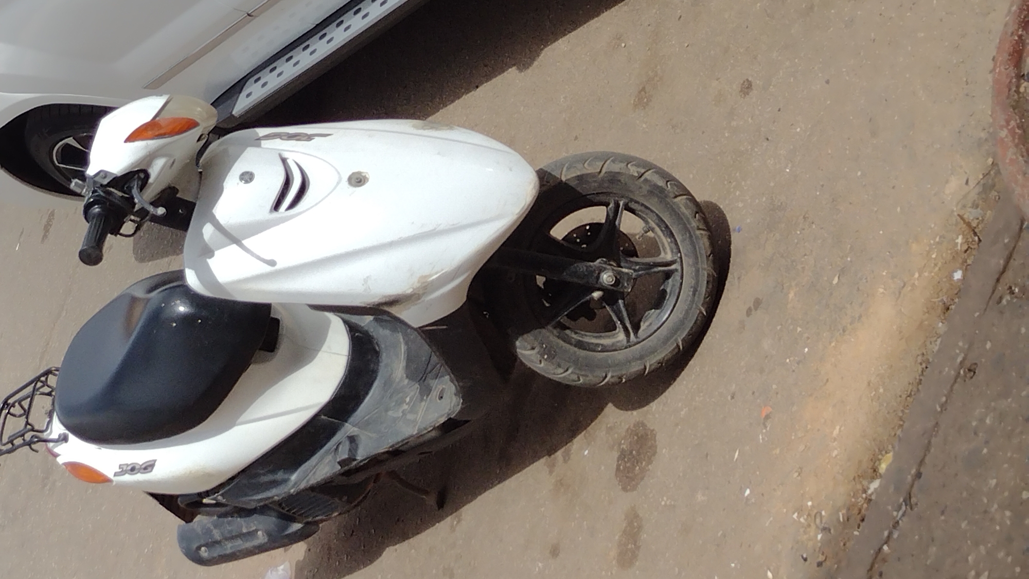 A slightly elevated shot reveals a white Yamaha Jog scooter parked on a paved, dirt-streaked surface in Jos, Nigeria. The scooter, angled with its front wheel pointing upwards, is positioned next to a white car with a visible side step. The scooter's seat is black, and the body is marked with the "JOG" logo. A brown concrete wall borders the scene on the right side. The background shows a worn surface with uneven texture and discoloration. No people are visible in the frame.