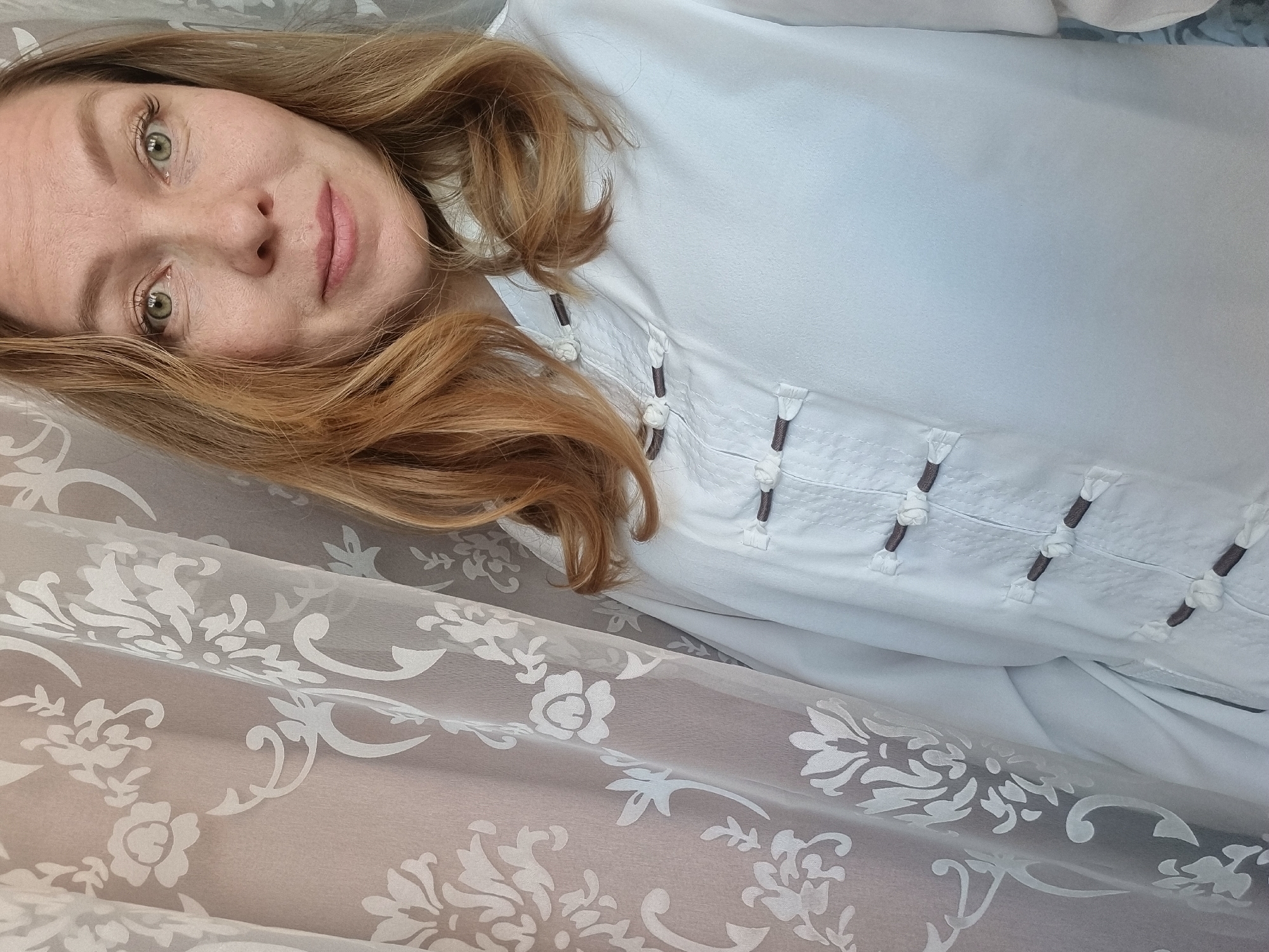 A woman with light-brown hair and green eyes is lying down, wearing a white blouse with decorative closures. She is positioned on a patterned fabric, likely a bedsheet or draped cloth. The scene is likely indoors, given the soft lighting and domestic setting. There is no explicit information indicating that the location is in Chaikovsky, Russia.