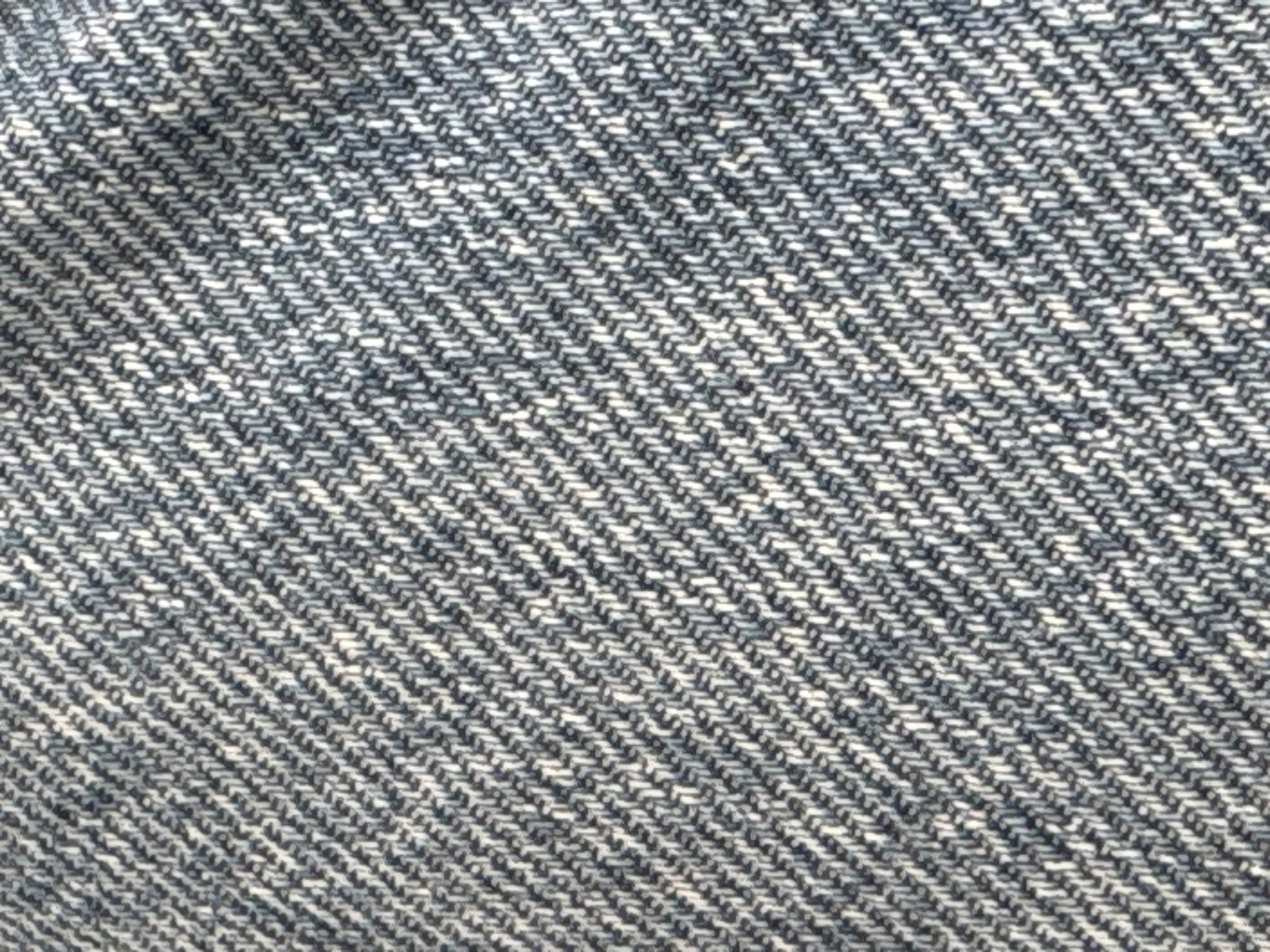 The image shows a close-up view of blue denim fabric. The texture of the denim is visible, with the characteristic diagonal weave pattern of twill. The threads are a mix of dark blue and lighter blue or white, creating a speckled or heathered effect. The lighting appears to be natural, casting subtle shadows that emphasize the texture of the fabric. There are no people, objects, or environmental elements present other than the fabric itself. The scene is entirely focused on the detail and texture of the denim.