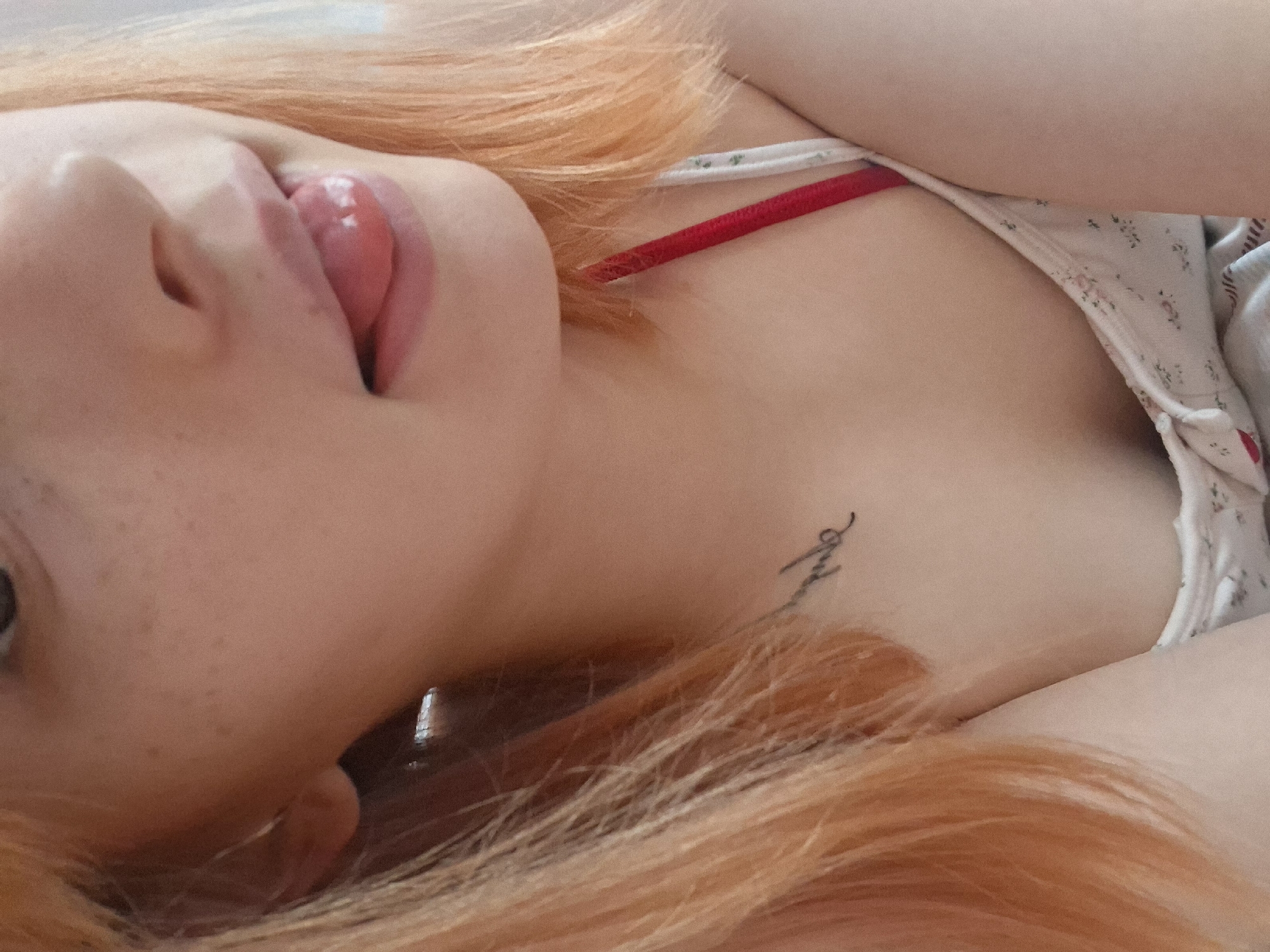 A fair-skinned individual with red hair is lying down, viewed from a low angle. The person is sticking their tongue out. They are wearing a white top with small floral prints and a visible red strap. A small tattoo is visible on their chest. The location is not discernible from the image.