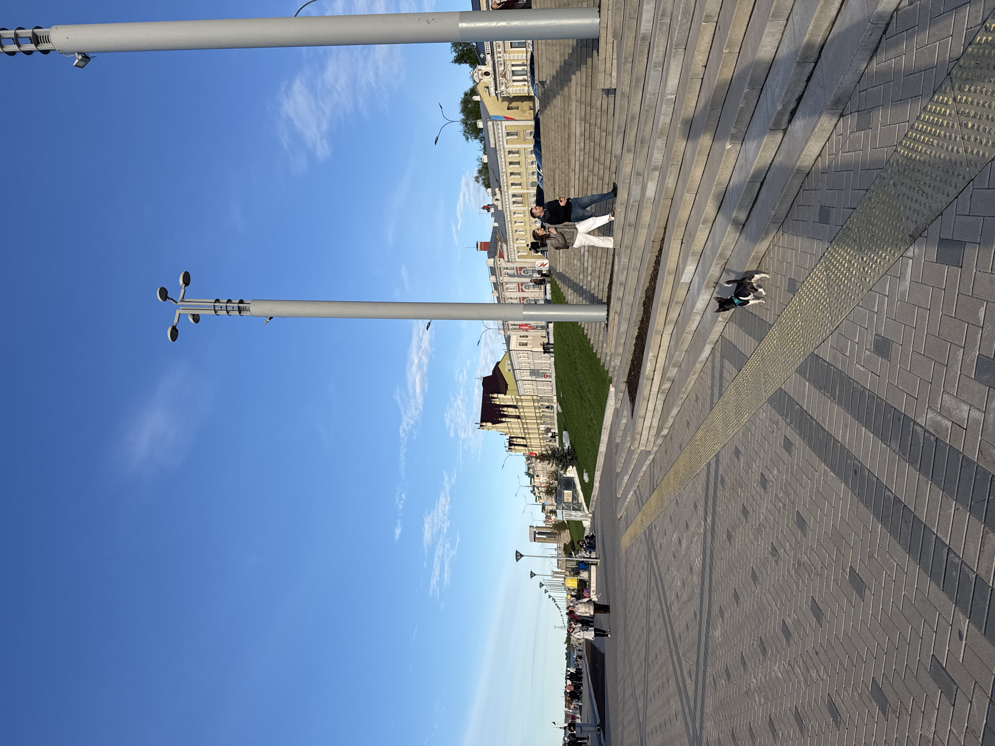 A ground-level view captures an outdoor scene in Nizhny Novgorod, Russia. The bright blue sky with wisps of clouds contrasts with the gray pavement in the foreground. The scene is dominated by gray brick, which is horizontally oriented in the picture. A yellow textured safety strip runs along the edge of the pavement. Two parallel silver light posts rise vertically into the sky.
On a set of gray stairs sit a man and a woman, while a dog stands nearby. Adjacent to the stairs is a strip of manicured grass, followed by buildings of varying heights. The scene extends into the distance. Silhouetted figures can be seen walking along a path further away.
