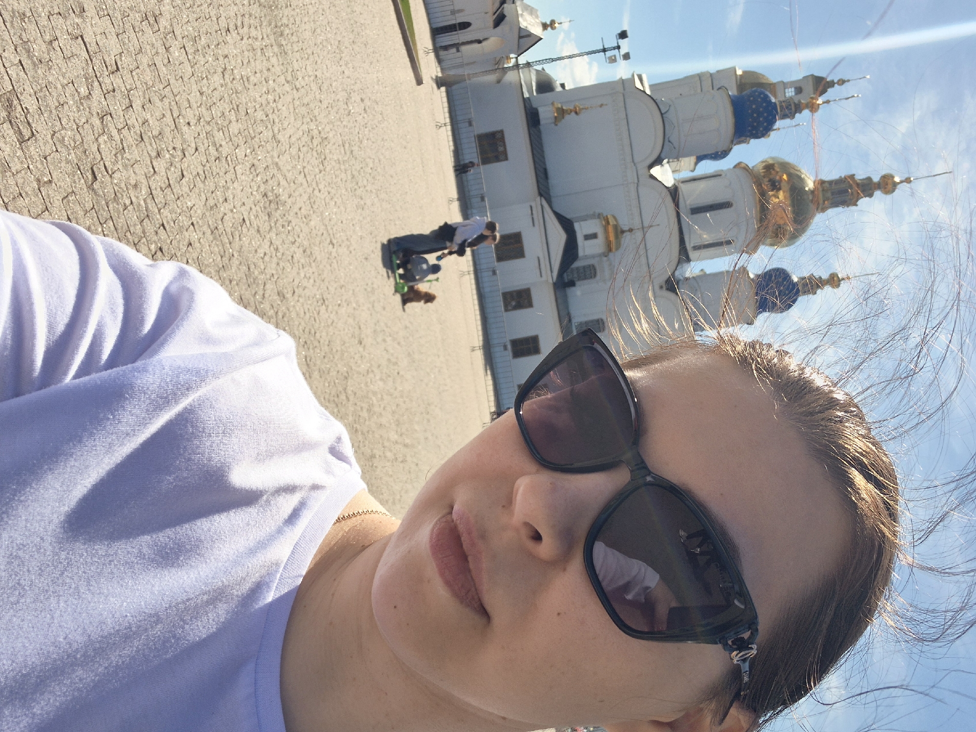 A selfie taken in Tobolsk, Russia. A woman with sunglasses, wearing a pale lavender shirt, is the foreground subject. In the background are the towers of the Tobolsk Kremlin's Saint Sophia-Assumption Cathedral. Further in the background a man with two children on a scooter, and a small dog. The ground is paved with rectangular stones. The sky is bright with light clouds.