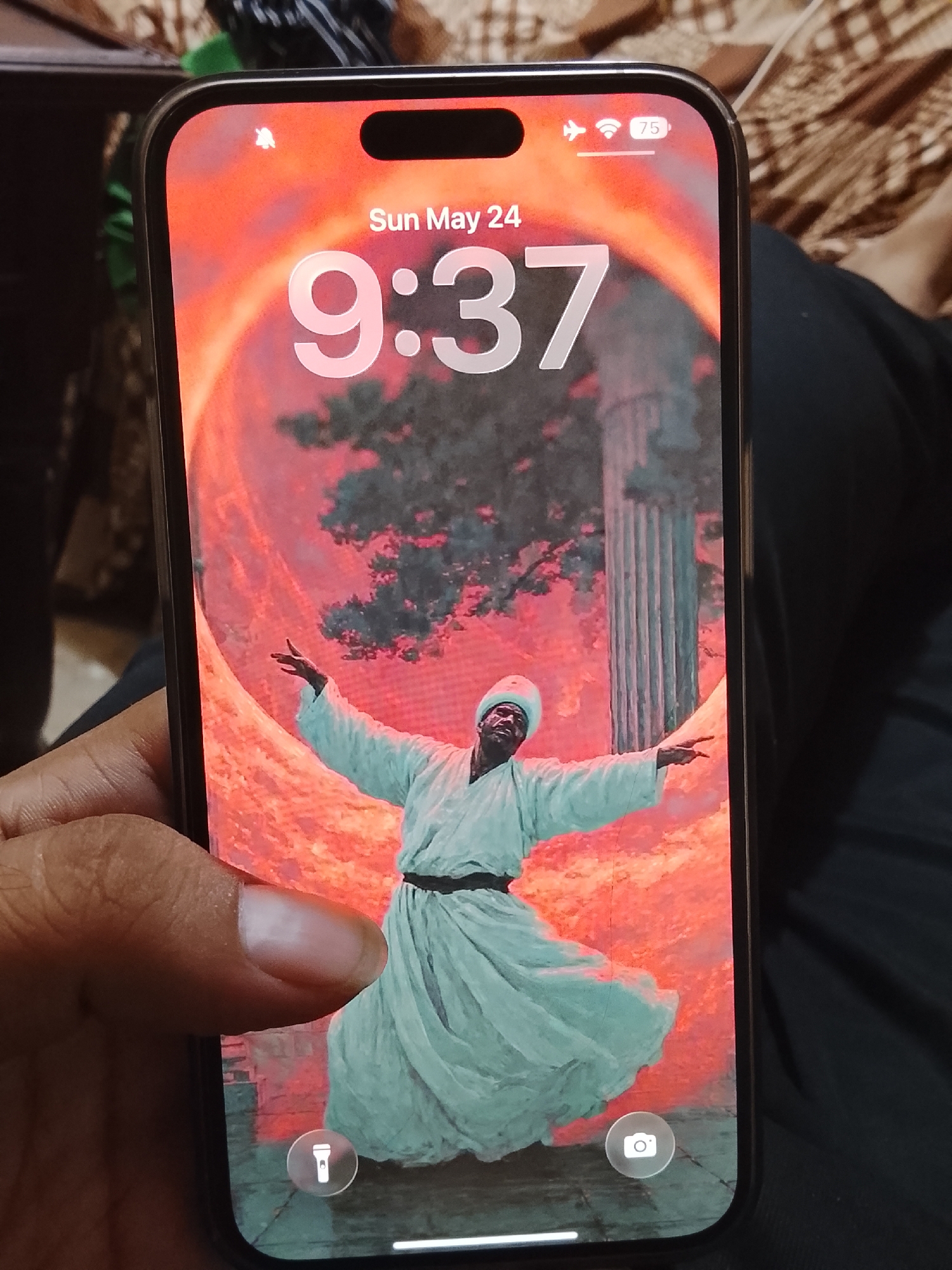 A hand is holding a smartphone displaying a lock screen with the time 9:37 AM and the date Sun May 24. Notification, signal, and battery indicators are visible at the top of the screen. The background wallpaper is an image of a man in a white robe and hat with arms outstretched. The setting of the wallpaper appears to be an open-air location with a tree and a column in the background, overlaid with red hues. The phone features a flashlight and camera icon at the bottom of the screen. The background around the phone features a dark garment. There are no indicators about the city of Multan, Pakistan.