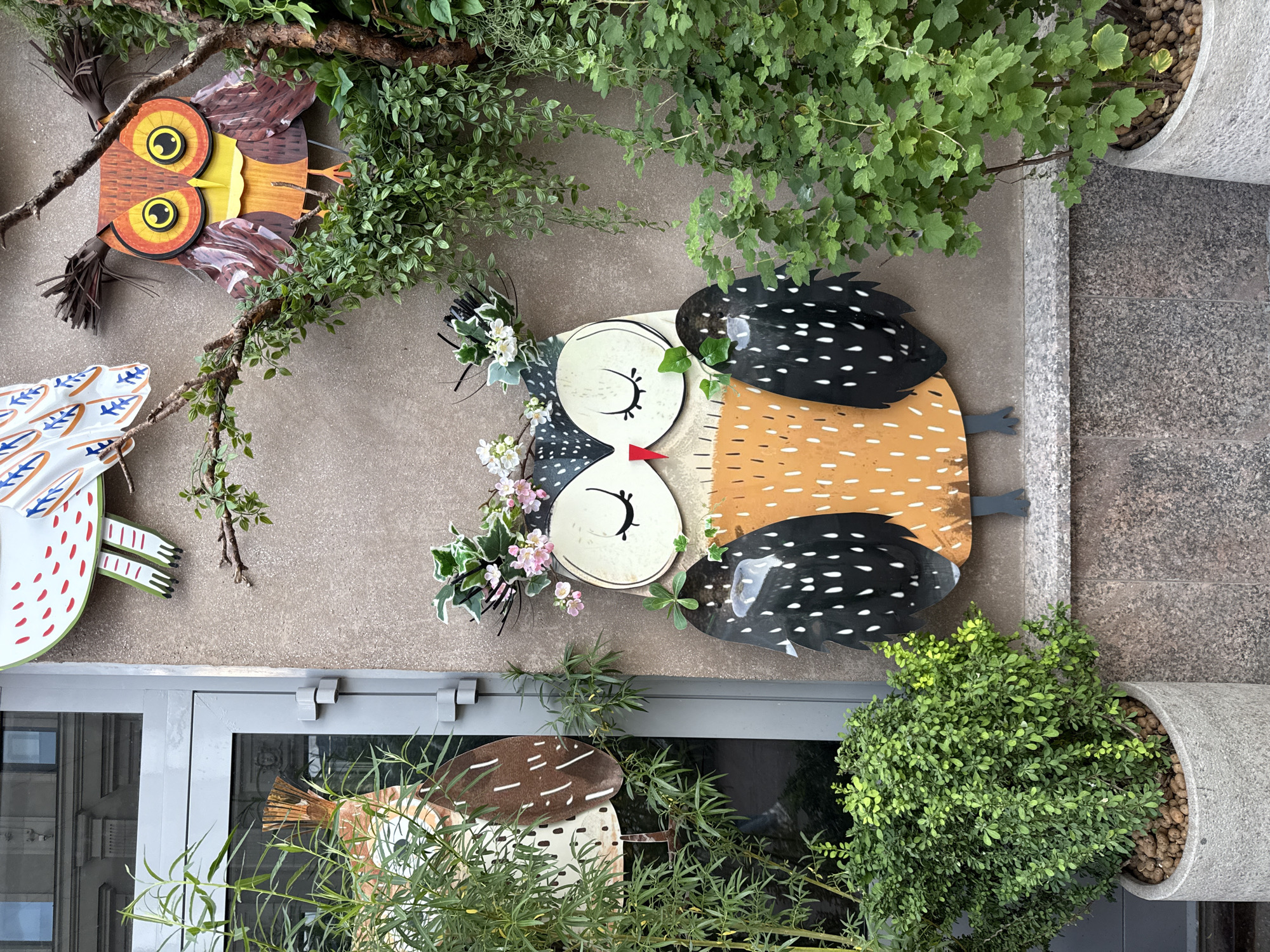 This media file captures a decorative outdoor display featuring three whimsical owl figures and various types of greenery, mounted on a building facade. The image is taken in a portrait orientation, with the display appearing rotated 90 degrees clockwise.
In the upper part of the frame, a brown and orange owl with large yellow eyes and black pupils is visible, its body textured to resemble feathers. Below and slightly to its right is a larger, more stylized owl. This owl has a cream-colored body with dark speckled patterns, closed eyes with prominent black lashes, and a small red beak. Its black wings are dotted with white spots, and its head is adorned with artificial pink and white flowers and green foliage. Partially visible at the bottom left is a third owl, primarily white with red and blue markings and green feet.
The owls are set against a light brown or beige textured wall. Interspersed among the figures are various real and artificial plants, including leafy vines and branches. To the right, a grey concrete planter holds a vibrant green bush with small leaves, the top of the soil covered in brown pebbles. Another potted plant with thin green leaves is visible at the bottom.
Below the main wall, a grey architectural element, possibly a window or door frame, reflects parts of a city scene, including other buildings. To the far right, a darker grey, speckled granite-like surface can be seen. The lighting is bright and even, suggesting daytime with either overcast conditions or indirect sunlight. The overall impression is decorative, cheerful, and artistic. No text is visible in the image. The location context provided states this is in Moscow, Russia.