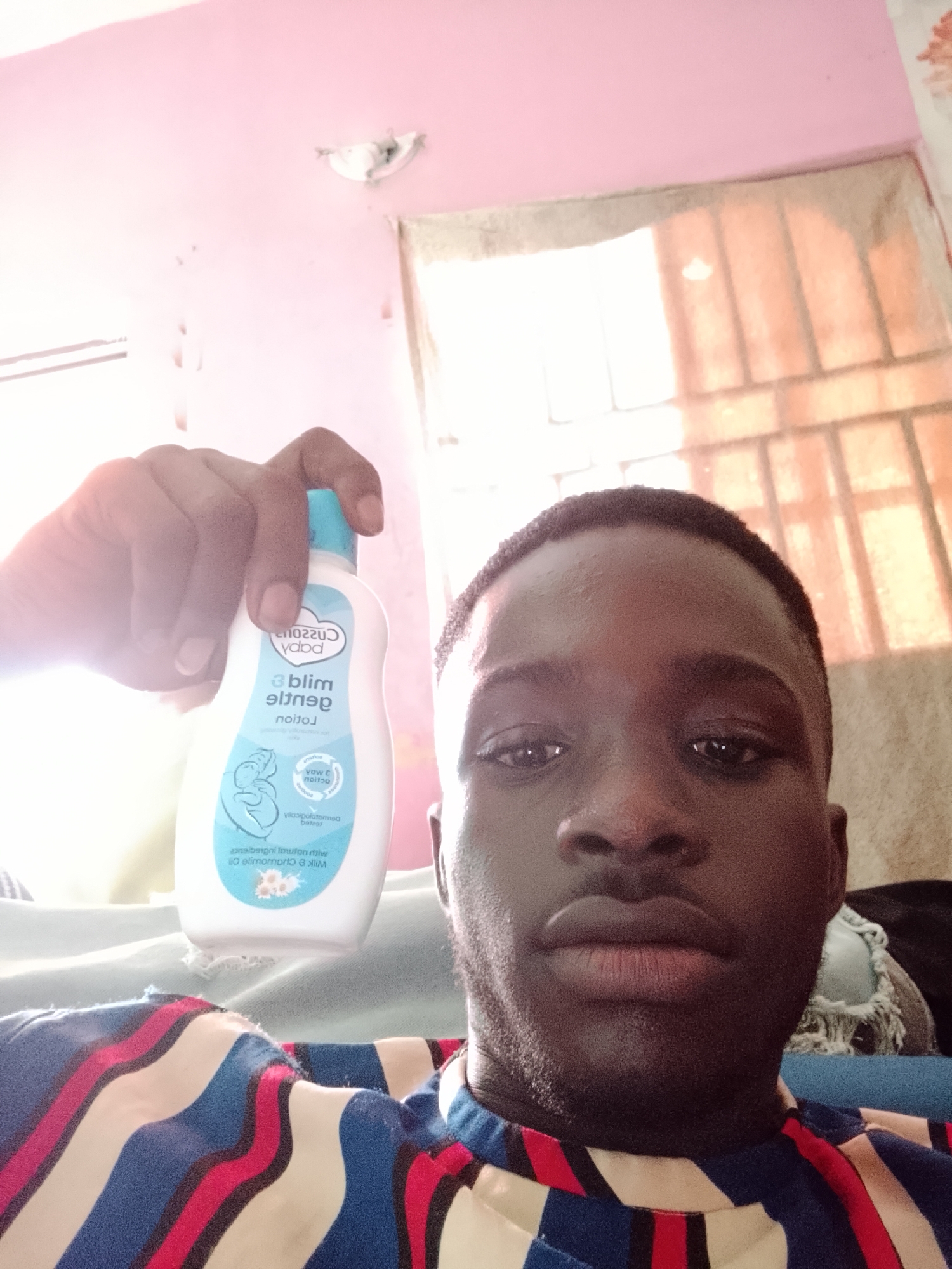 This media file is a selfie taken indoors. The primary subject is a young man with dark skin and short hair, looking directly at the camera. He is wearing a colorful striped shirt with bold bands of blue, white, red, and yellow. In his right hand, held up towards the camera, is a white bottle with a light blue cap. The bottle has text and graphics printed on it, including the words "baby mild gentle lotion" and an illustration of a person holding a baby. The background of the image is somewhat blurred but appears to be a room with pink walls and a window with blinds. The lighting suggests it is daytime, possibly with natural light coming from the window. The overall mood appears casual and personal. The text on the bottle, though partially obscured and reversed, indicates it is a baby lotion. Given the location context of Ihiagwa, Nigeria, this is likely a personal photograph taken within a home.