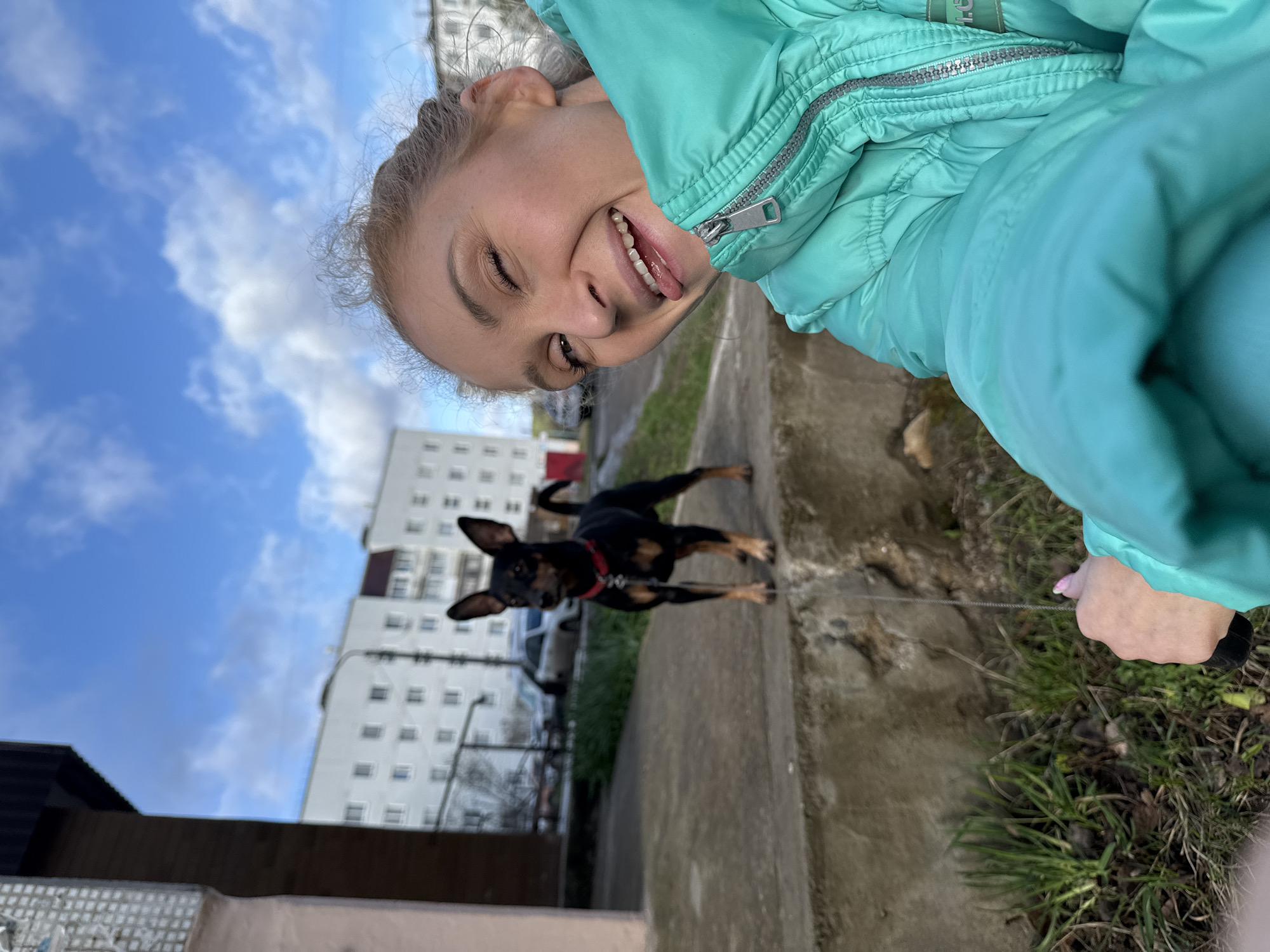 A cheerful outdoor selfie captures a young woman and her dog on a walk in Chaikovsky, Russia. The woman's face, positioned in the upper right of the frame, beams with a wide smile, her tongue playfully peeking out. She wears a vibrant teal puffer jacket, its silver zipper visible, suggesting cooler weather. Her hand, adorned with light pink nail polish, holds a thin leash.
The leash extends to a small, black and tan dog with erect ears and a red collar, captured in motion on a concrete path. The dog appears to be a Miniature Pinscher or similar breed, enthusiastically moving forward. In the background, tall, white multi-story apartment buildings with darker accents stand under a bright blue sky scattered with white clouds. Patches of dry grass and dirt border the concrete path. The natural light suggests it is daytime. The low-angle shot and the woman's expression convey a sense of joy and playful interaction during a casual stroll with her pet in an urban setting.