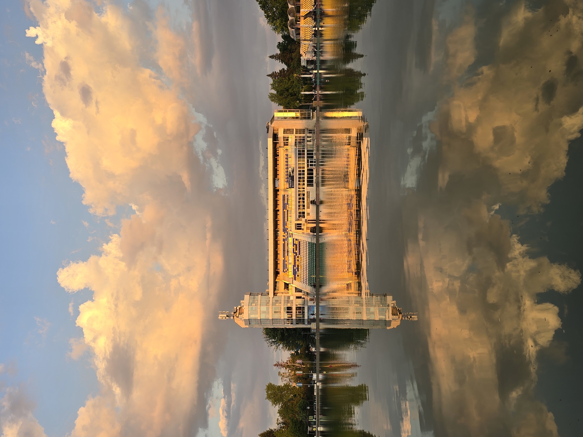 The image depicts a building reflected in water. The building is multi-story with a modern design, featuring a predominantly glass facade and a lighter-colored exterior. Trees and another structure are visible on the far side of the water. The sky is filled with clouds that are reflecting in the water along with the building. The colors of the image are primarily warm tones, with orange and yellow hues dominating the sky and the reflection in the water.
The location is Unknown, Unknown.