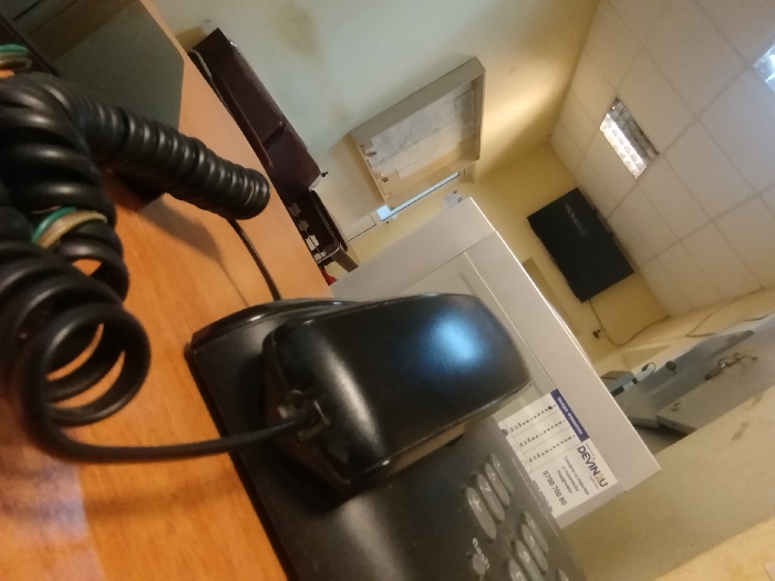 The scene is an interior view of an office in Boychinovtsi, Bulgaria, photographed from an oblique, elevated angle. A black landline telephone handset rests on its base on a light-brown wooden desk in the foreground. Its coiled cord is visible on the desk surface. Adjacent to the telephone is a light-colored office machine featuring a label with "DEVIN.U" and a service number "0700 700 80". The room has light-yellow walls and a suspended ceiling with rectangular panels, one of which contains an illuminated fluorescent light fixture. In the background, a dark wooden cabinet stands against a wall, and a black flat-screen monitor or television is wall-mounted. No persons are present. The area is illuminated by overhead artificial lighting.