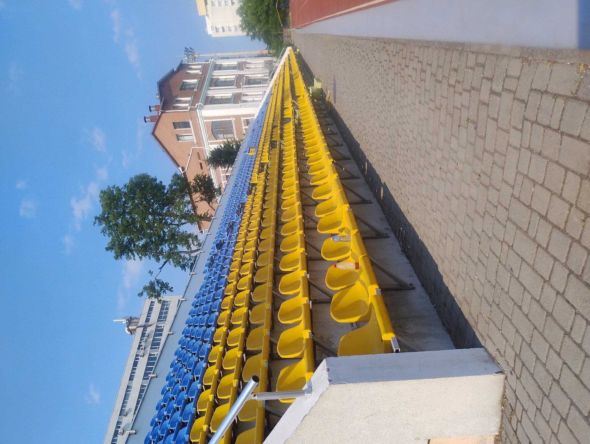 An outdoor, high-angle, eye-level shot shows rows of empty stadium seats. The seats are blue and yellow and are arranged in long rows. A paved walkway runs alongside the seats. In the background, there are several buildings and trees. The sky is clear and blue. The sun appears to be shining, casting shadows on the walkway and seats. The scene appears to be a sports stadium or arena. No people are visible in the image, and no text is present. The time of day appears to be daytime, likely late morning or early afternoon based on the sun's angle and the shadows. The weather appears to be clear and sunny. The location is an outdoor stadium.