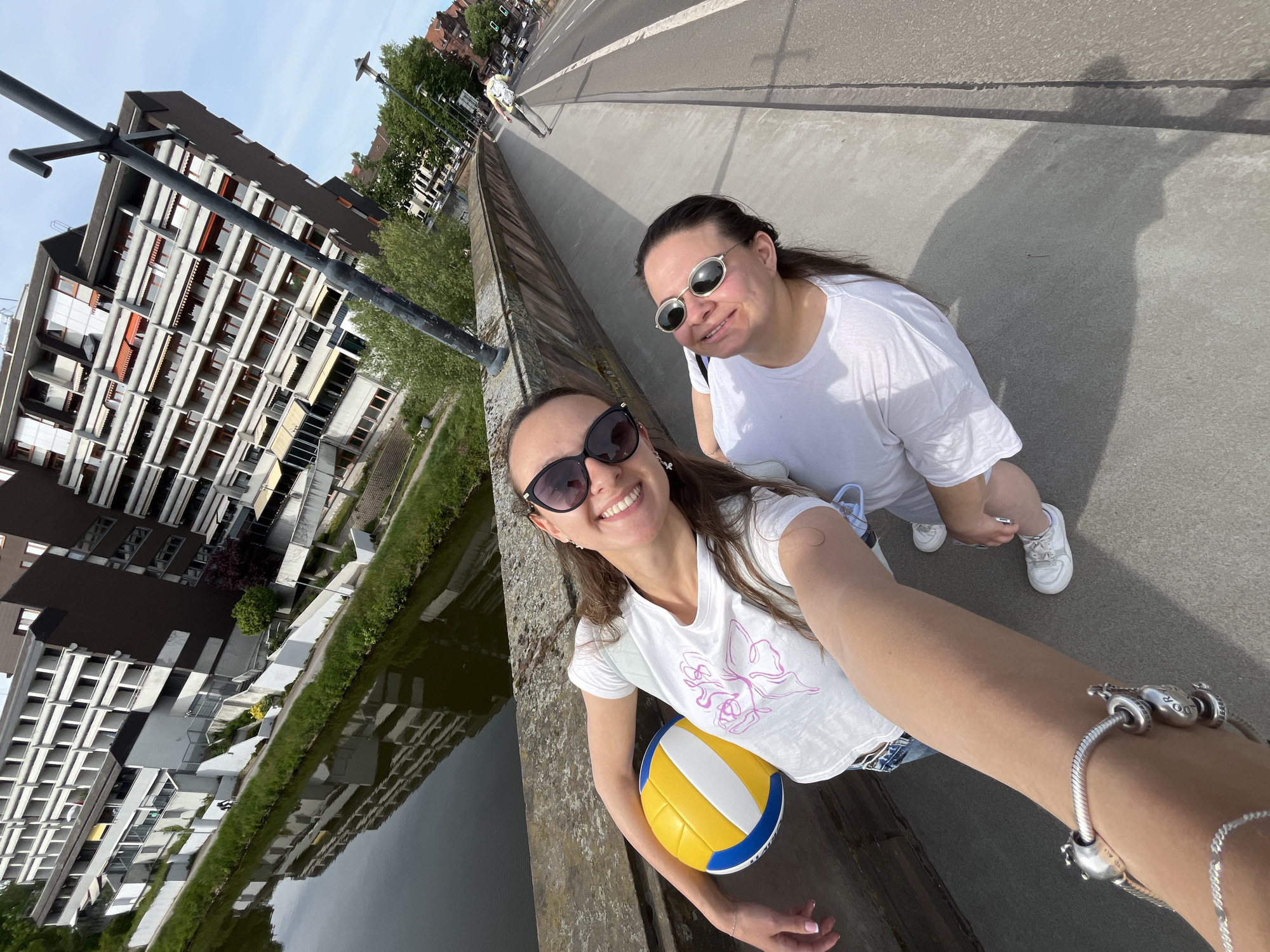 Two women pose for a wide-angle selfie on a bridge in Saarbrücken, Germany, overlooking an urban canal or river. Both women are smiling and wearing sunglasses, indicating a bright, sunny day. The woman in the foreground, on the left, has long brown hair and wears black sunglasses, a white t-shirt with a pink graphic design (possibly a butterfly or abstract shape), and holds a yellow, blue, and white volleyball in her left arm. She also wears a silver charm bracelet on her right wrist. The second woman, behind and to the right, wears round, silver-rimmed sunglasses and a plain white t-shirt, kneeling slightly. Her shadow is visible on the concrete bridge surface, emphasizing the strong sunlight.
The setting includes a modern, multi-story building with numerous balconies reflected in the still water of the canal or river below the bridge. Lush green grass and trees line the banks of the waterway. The bridge itself features a concrete barrier, and a road is visible behind the women, with another person walking further down the road. The architecture of the visible buildings is contemporary, characterized by block-like structures and repetitive window patterns. The women appear to be enjoying their time outdoors. Part of the text "MR.BAL..." is visible on the volleyball.