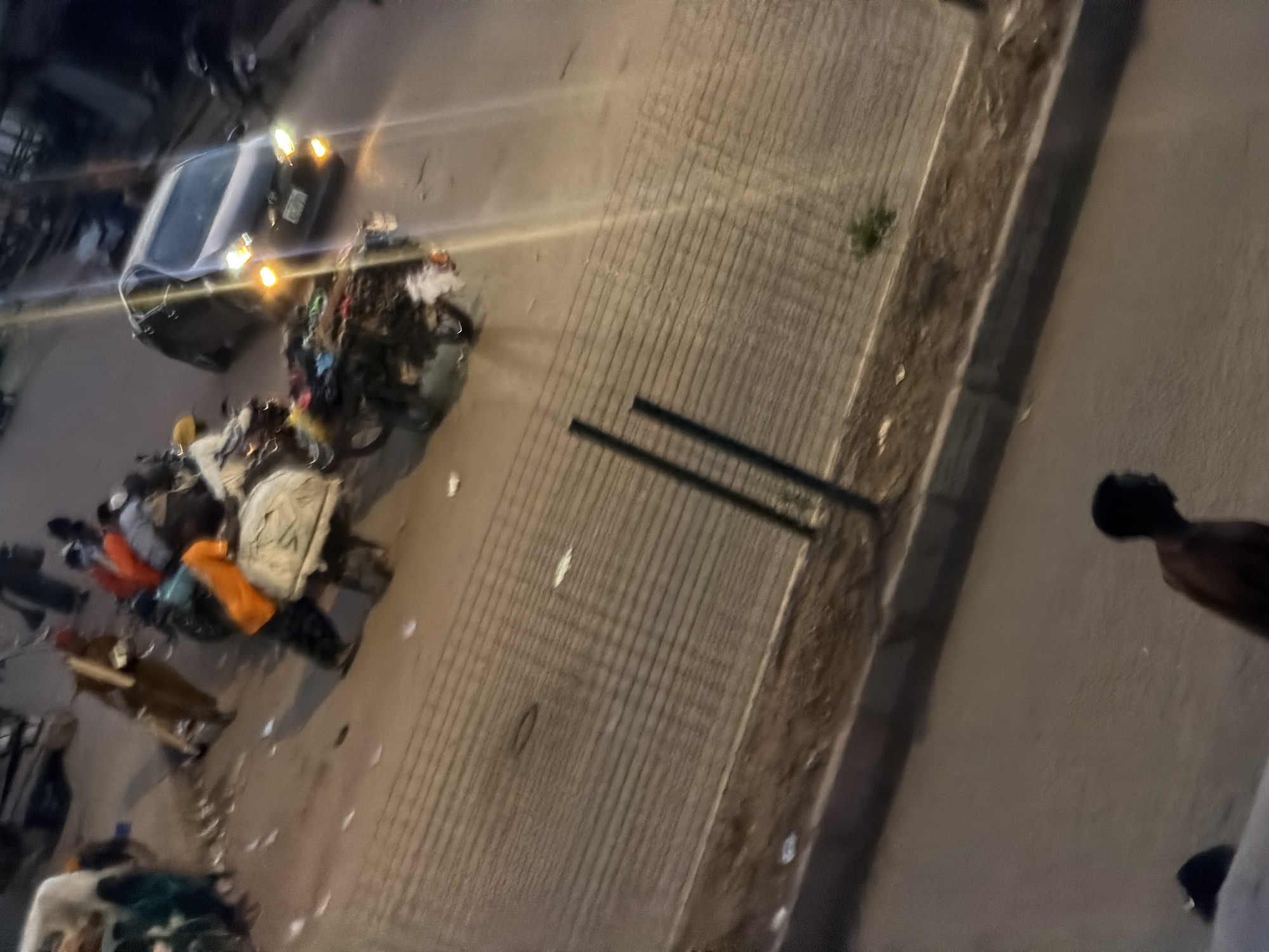 The image depicts a street scene in Bukuru, Nigeria, likely at dusk or nighttime, given the artificial lighting from vehicles and the overall dimness. In the foreground, a person's torso and head are visible from the shoulders up, facing to the right, with their back partially to the viewer. The person is bare-chested.
Further back on the road, a dark-colored sedan with its headlights and hazard lights illuminated is in motion. Adjacent to the car, a motorcycle is laden with a large, overflowing pile of goods, appearing to be clothing or textiles, possibly being transported. Several people are clustered around this motorcycle and in the vicinity, some appearing to be loading or securing the cargo. One individual is wearing an orange garment. Another person is visible in the background, walking with a tan-colored object, possibly a broom or stick, over their shoulder.
The street appears to be paved and is lined with a textured barrier or wall, possibly a sidewalk or a drainage channel, with a wire mesh fence or railing running along its top edge. Scattered debris, including small white objects, are visible on the road surface. The environment suggests a busy urban setting, possibly a market or transport hub, where goods are being moved and people are engaged in various activities.