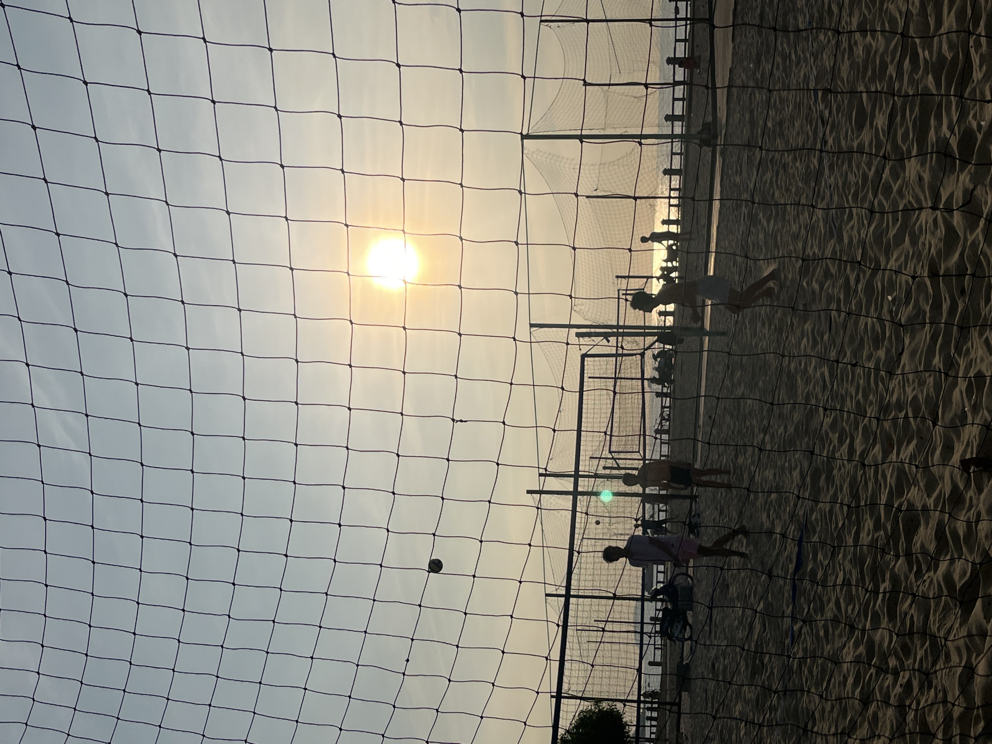 The photograph depicts a beach volleyball game in Nha Trang Ward, Vietnam, viewed through a foreground volleyball net. The sun is visible in the sky, causing a silhouetted effect on the players and the beach environment. Players are actively engaged in the game. A volleyball is mid-air. Other nets are in the background. The beach is sandy.