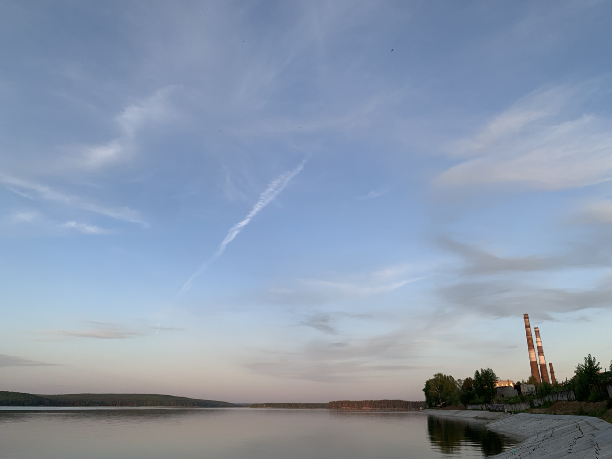 The image depicts a calm body of water with forested land in the distance. The sky is predominantly blue with scattered clouds and a contrail. On the right side of the frame, there are industrial structures, including two tall smokestacks with red and white stripes. These structures are positioned near a concrete embankment and vegetation. The image is likely taken near a body of water in or near the city of Chaikovsky, Russia, given the presence of industrial architecture typical of the area. No people or specific actions are discernible in the scene. The environment suggests a late afternoon or early evening time due to the soft lighting.