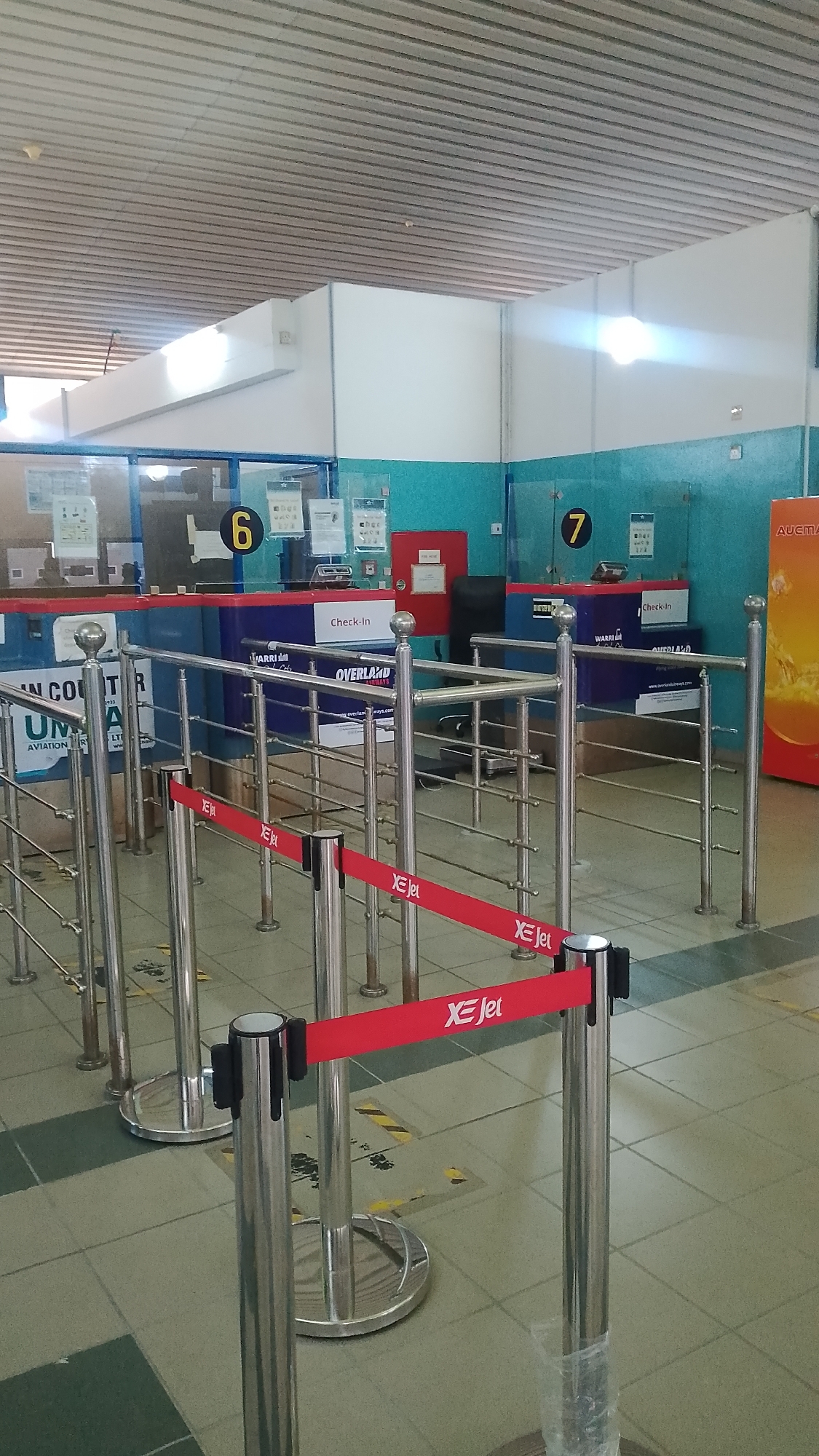 The image depicts the interior of an airport terminal, specifically the check-in area. Several queuing stanchions with red retractable belts labeled "XE Jet" are set up, leading to check-in counters numbered 6 and 7. The check-in counters are for Overland Airways, and a sign with "WARRI" on it is visible on each counter. The walls behind the counters are painted blue and have signage posted on them. The floor is tiled in a light green and grey pattern, with some yellow and black caution tape on the floor. An "AUCMA" advertisement is visible on the right side of the image. The ceiling is white and features recessed lighting. The location is likely in Orerokpe, Nigeria, given the text on the image and the presence of Overland Airways, a Nigerian airline. There are no people visible in the frame.