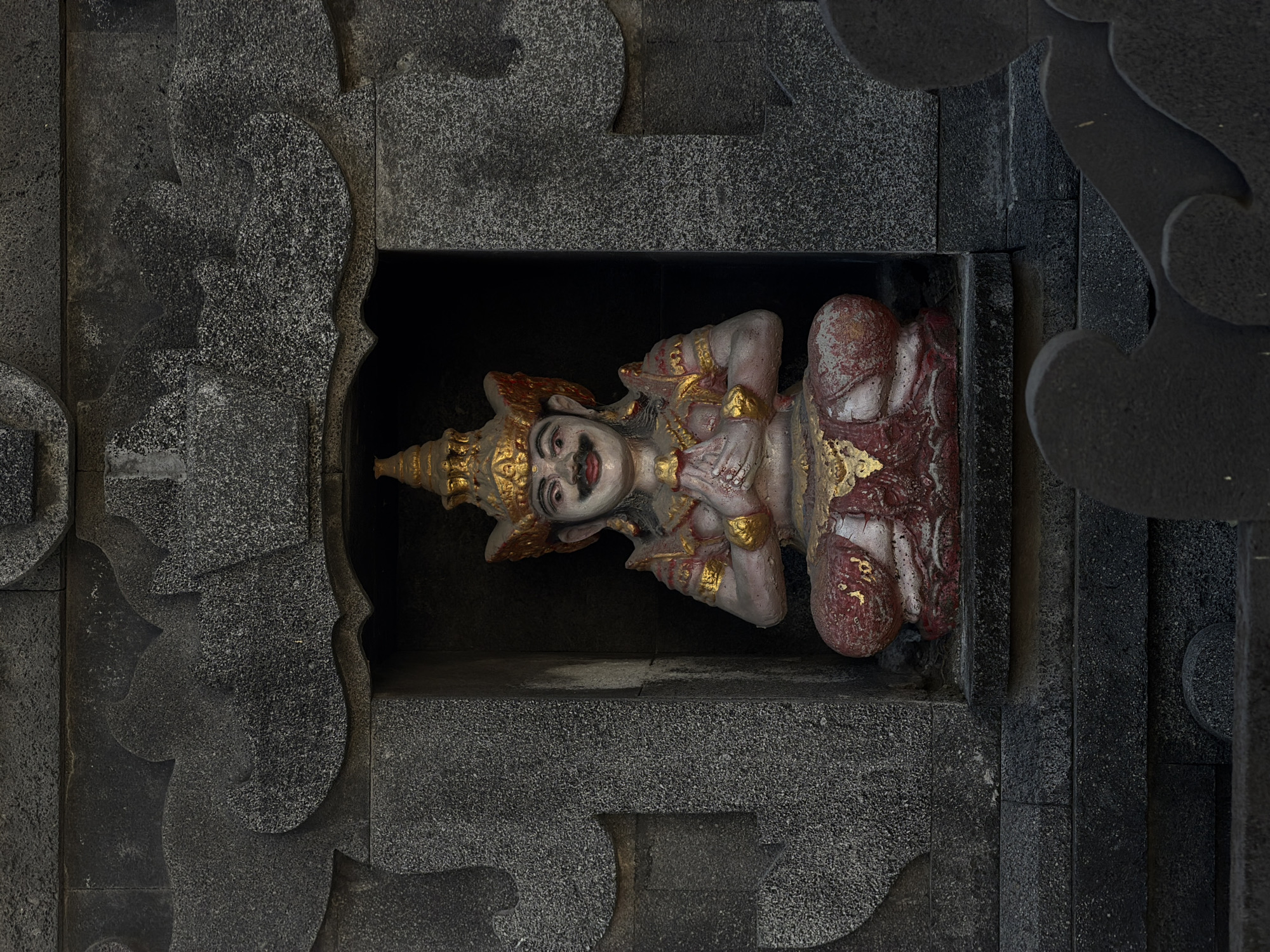 A stone statue of a deity with a smiling face, adorned with a golden crown, bracelets, and other ornaments, sits in a rectangular alcove of a building in Mengwi, Indonesia. The statue is painted in white, red, and gold colors. The surrounding alcove is made of dark gray stone. The alcove is intricately carved with curved, layered designs. The statue appears to be a religious figure or guardian spirit, likely a part of a local Hindu or Buddhist temple. The setting appears to be an old building, possibly a temple or public building.