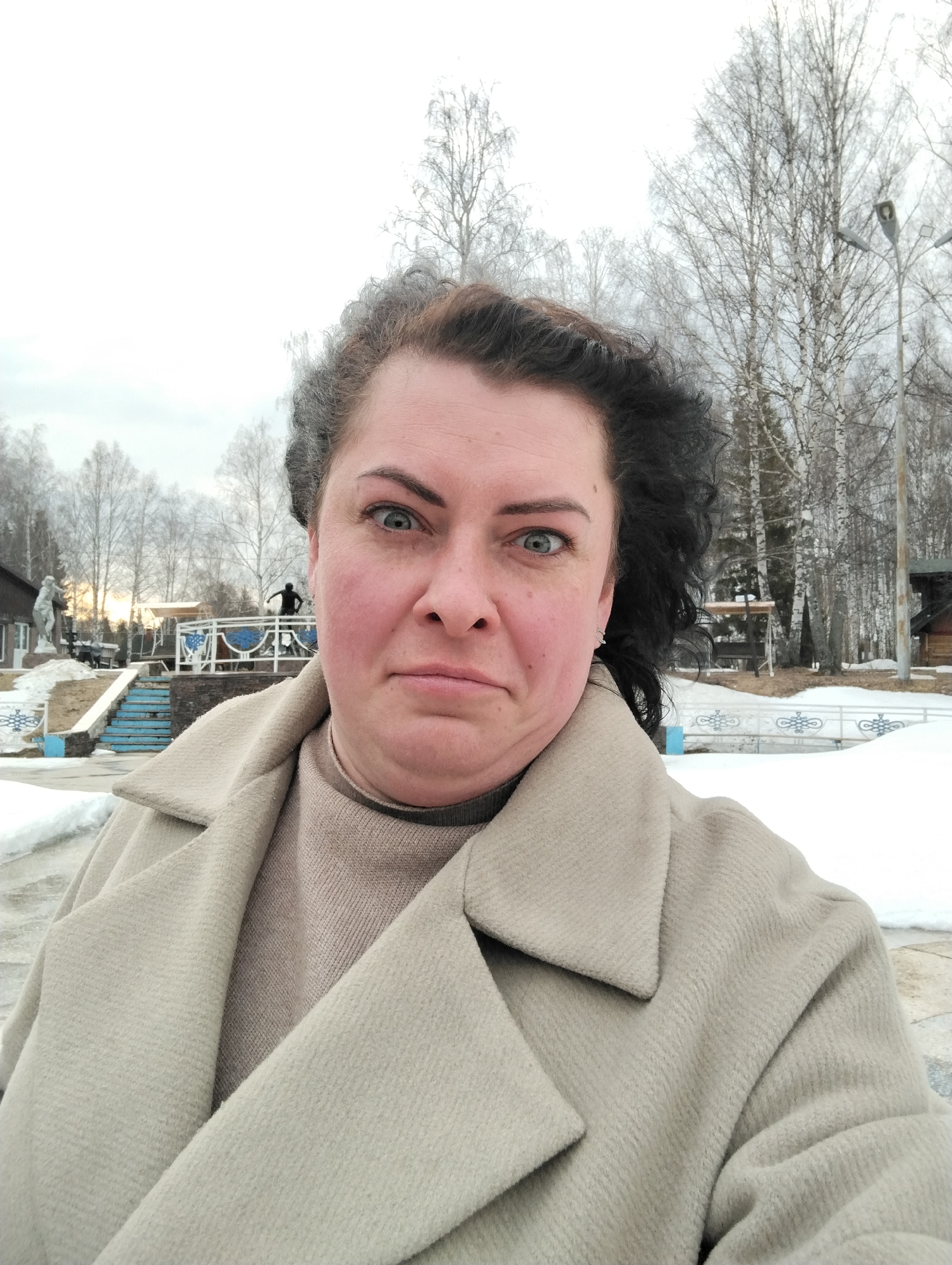 A medium-shot selfie shows a woman outdoors in Galevo, Russia. She has gray-tinged brown curly hair, fair skin with flushed cheeks, and blue eyes. She wears a beige coat and a beige turtleneck sweater. In the background is a park scene, partially covered in snow. Trees with bare branches are visible, along with a building, statues, and a blue-and-white railing. A figure in dark clothing stands on a raised platform. The sky is overcast and pale.