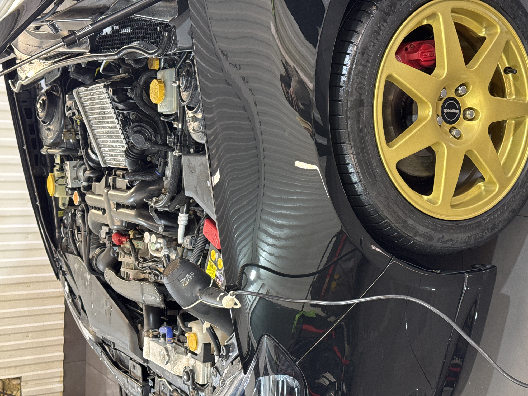 The image shows the front engine compartment and front passenger-side wheel of a black car. The engine appears to be a horizontally opposed four-cylinder (boxer) type, with a front-mounted intercooler. The wheel is gold-colored with a six-spoke design. The brake caliper is red and appears to have "STI" printed on it. A power cord is plugged into a device near the front of the engine compartment. The location is not discernible from the image, so it's impossible to confirm that it is in Chaikovsky, Russia.