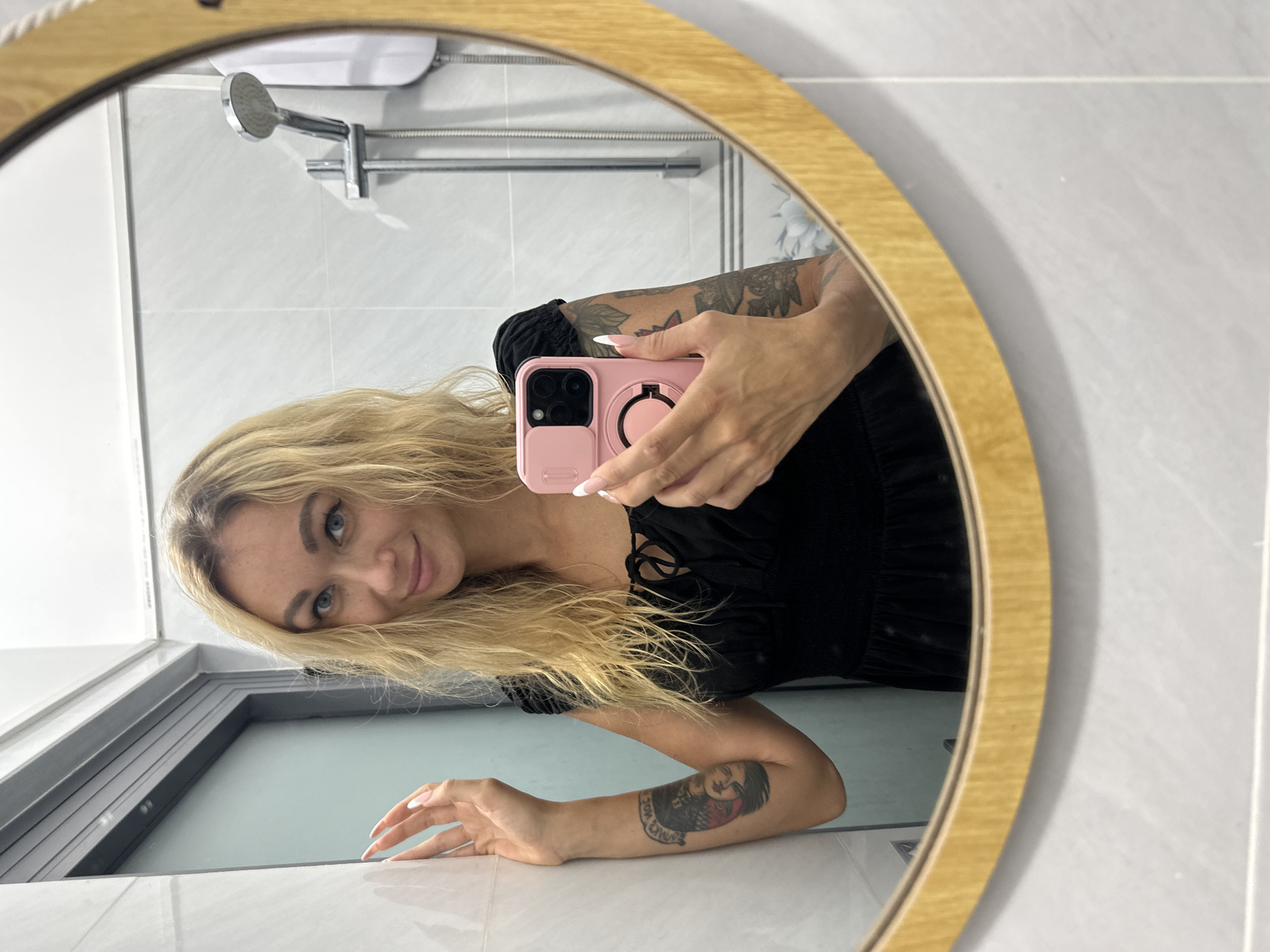 A blonde woman with wavy hair and light eyes is captured in a mirror selfie, taken in what appears to be a bathroom in Mũi Né, Vietnam. She is looking directly at the camera with a subtle smile. Her head is tilted, and her long blonde hair cascades over her shoulder and arm. She is wearing a black top or dress with ruffled sleeves.
She holds a pink smartphone in a matching pink case, featuring a circular ring holder on the back. Her hands have manicured nails with white tips. Tattoos are visible on both of her arms. The tattoo on her visible right forearm (her left arm) features abstract designs and what looks like partially legible text or numbers. The tattoo on her visible left forearm (her right arm) depicts a figure, possibly a woman's face, with some indistinguishable text below it.
The mirror she is using is round with a light wooden frame. Reflected in the background are light grey or white square tiles, typical of a bathroom wall. A silver shower head and an accompanying horizontal bar fixture are also visible in the reflection, indicating a shower area. Below the woman's face, a window-like opening or a low partition with a dark frame is visible, onto which she appears to be leaning with her left arm. The indoor lighting suggests it could be daytime or a well-lit interior. No clearly legible text is visible other than the faint, unreadable elements within her tattoos.