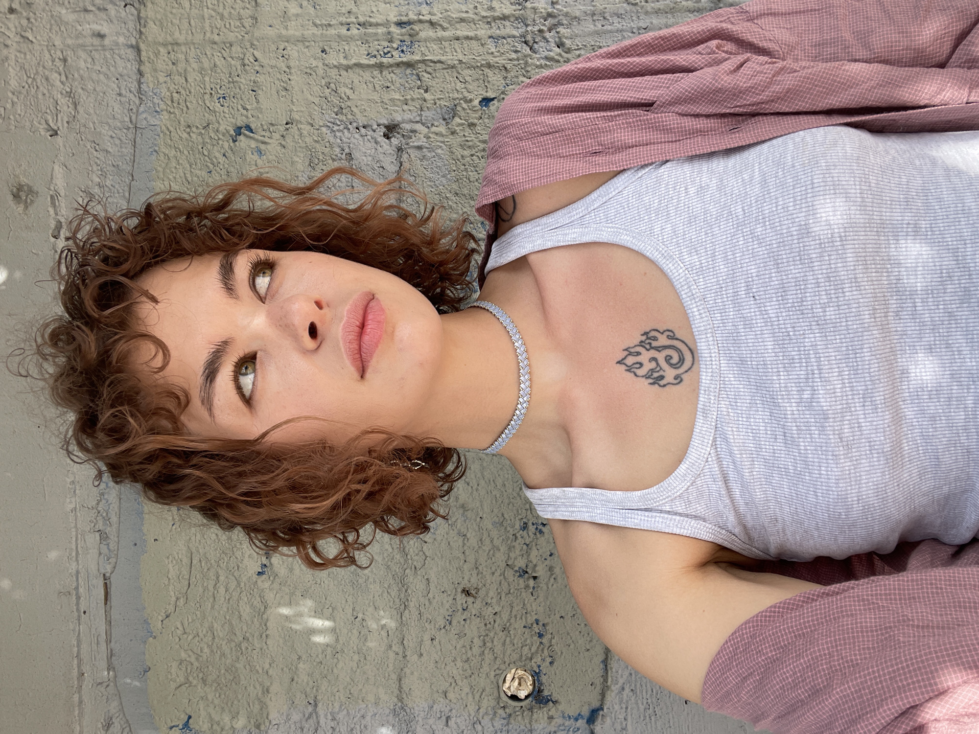 A young woman lies on a weathered, gray-painted concrete surface in Rafailovići, Montenegro. Her curly brown hair frames her face as she gazes upwards. She wears a light gray striped tank top and a pink plaid shirt. A delicate silver choker adorns her neck, and a tattoo of a cloud-like design is visible on her upper chest. The background shows a textured concrete wall with signs of aging and weathering.