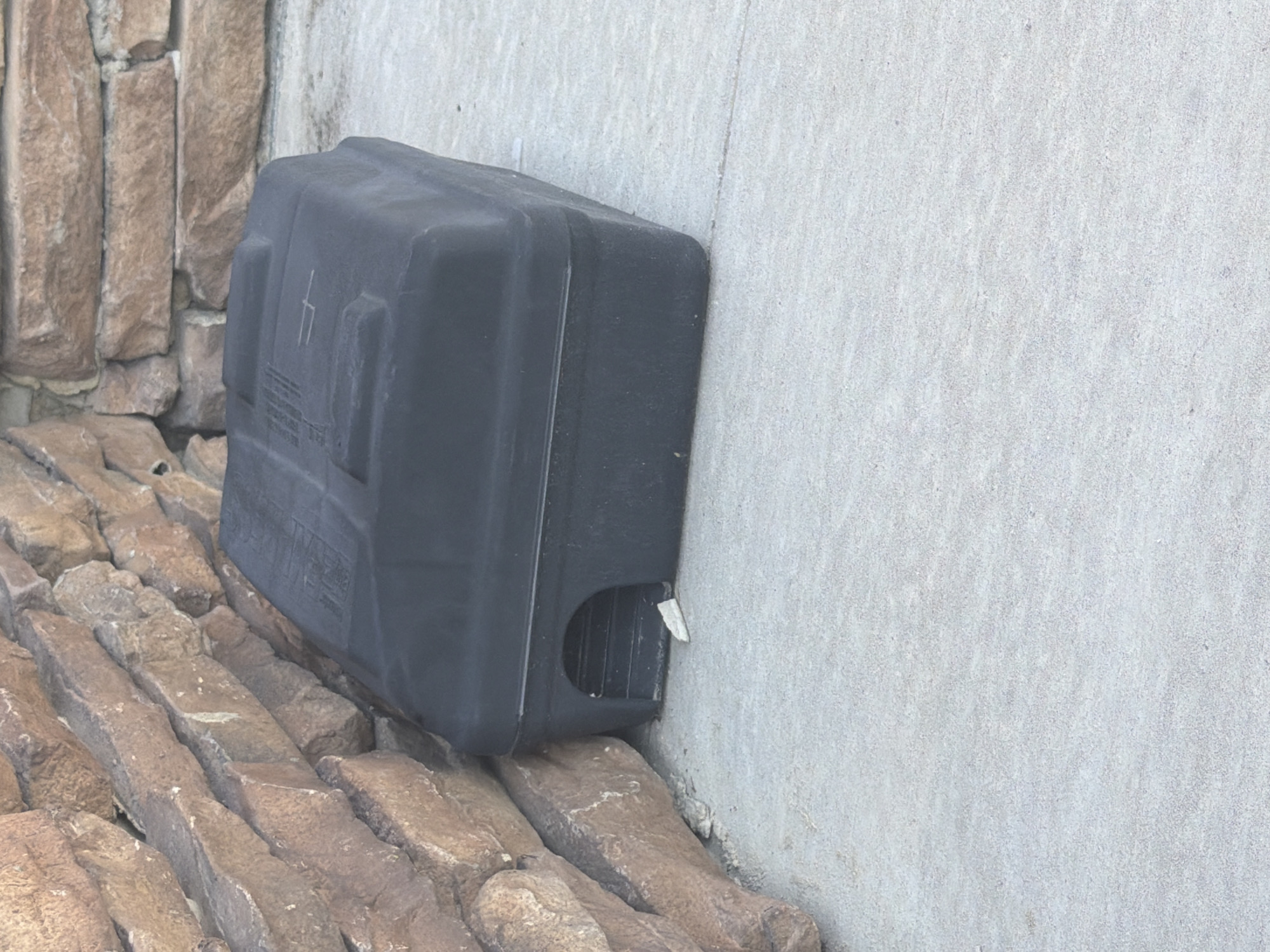 A black, rectangular bait station sits on a light gray concrete surface next to a stone wall. The bait station has a small, arched entryway on its left side. A small, white piece of material is visible just inside the entryway. The stone wall is constructed of irregular, earthy-toned stones. The scene appears to be outdoors in an urban environment.