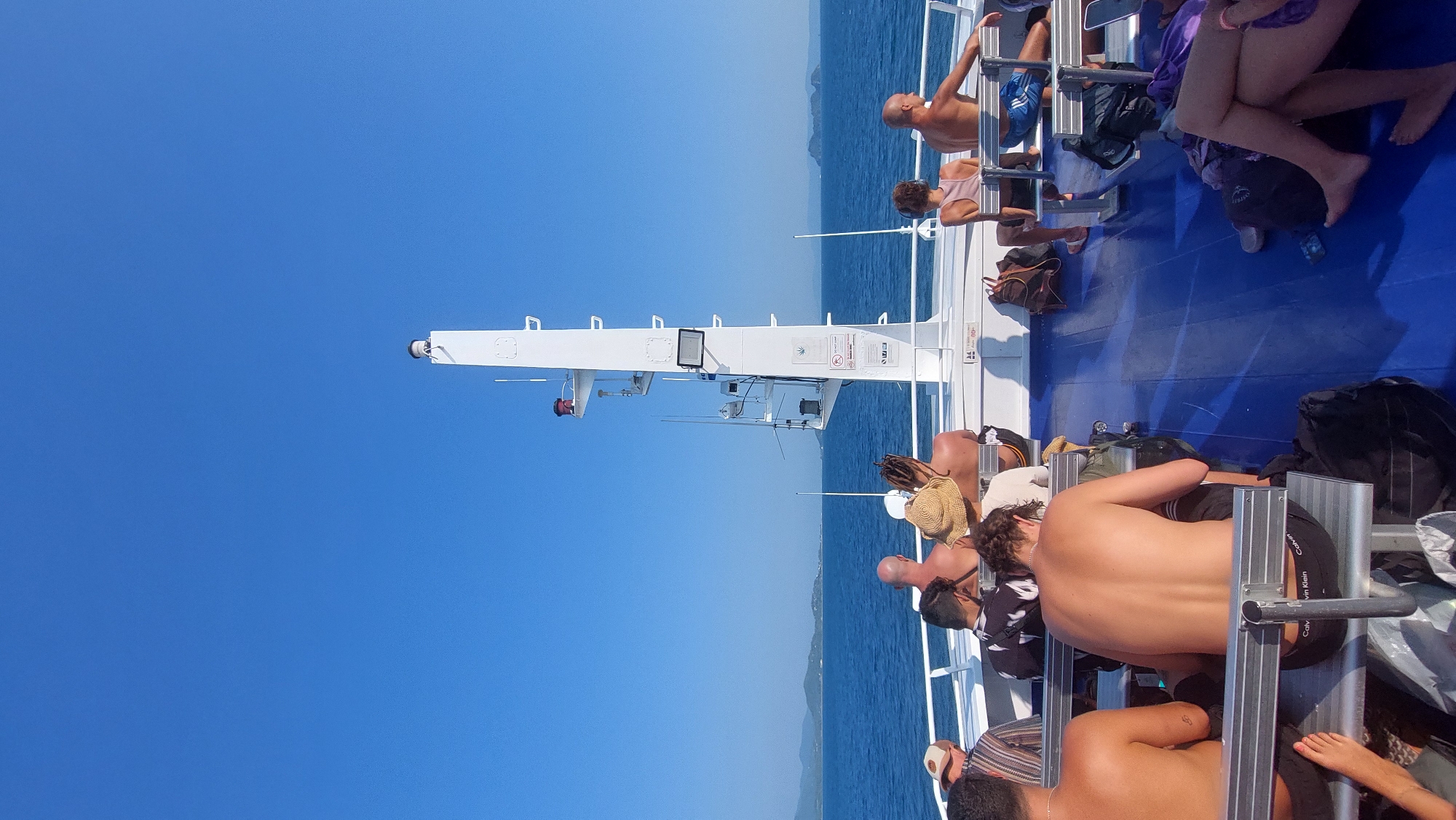 The image shows the deck of a ferry, crowded with passengers. A large white mast rises towards the clear blue sky. The sea is visible in the distance, with land on the horizon. Passengers are sitting or lounging on the deck. The location cannot be determined from the image as Baan Sri Thanu, Thailand.