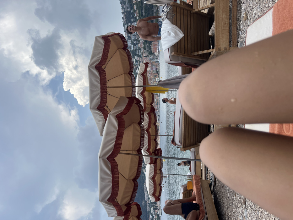 A beach scene features rows of matching fringed umbrellas. People lounge on beach chairs. A shirtless man stands near the umbrellas. Water and coastal buildings form a backdrop. In the foreground, a pair of legs are prominently displayed. The sky is visible with scattered clouds.
