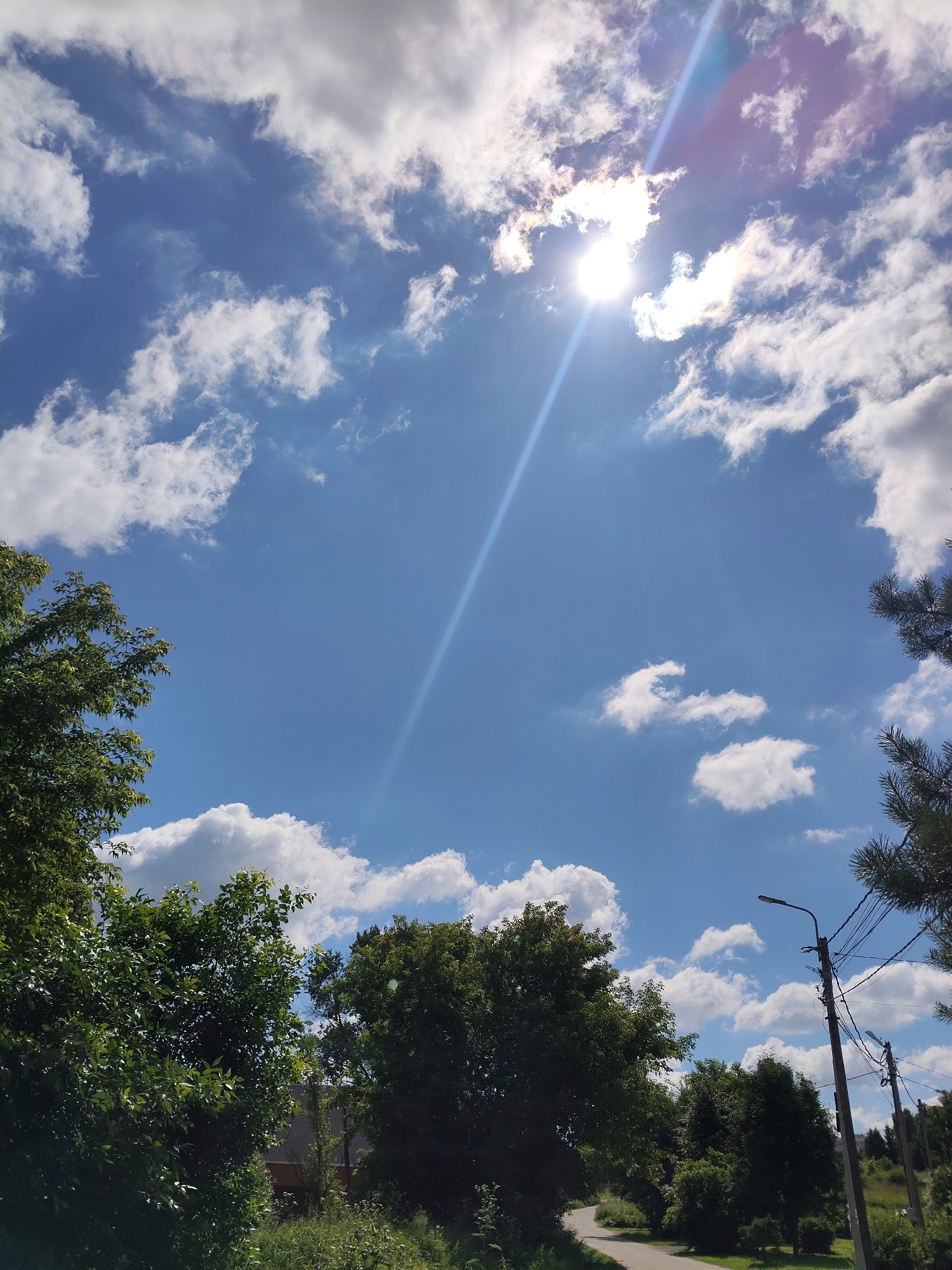 The image depicts a bright, sunny day in Venyukovo, Russia, with a clear blue sky filled with scattered white cumulus clouds. The sun is high in the sky, creating a lens flare that streaks across the upper portion of the image. In the foreground, lush green trees frame the scene on the left and right. A paved road curves through the landscape, leading into the distance. On the right side of the road, there are utility poles with wires and streetlights, suggesting a semi-urban or suburban setting. The overall impression is of a pleasant, natural environment on a warm day.