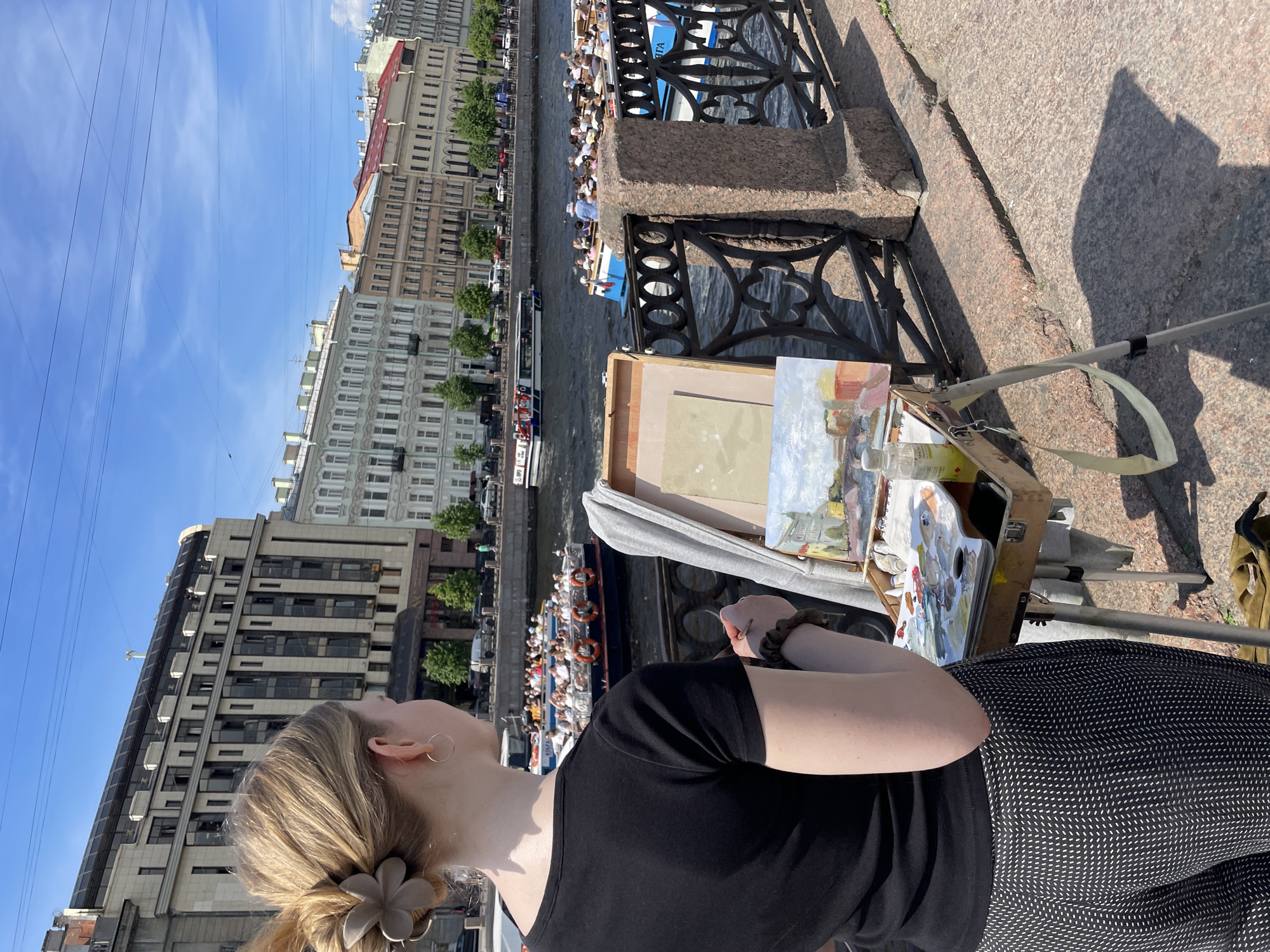 A young woman with blonde hair tied back in a bun is standing with her back to the viewer, painting a scene of a canal in Saint Petersburg, Russia. She is wearing a black t-shirt and patterned pants. In front of her is an easel holding a partially completed painting of buildings and boats on a waterway. Various art supplies, including tubes of paint and a palette, are arranged on a wooden box beneath the easel. The canal is lined with ornate buildings and a bridge with a decorative metal railing. Several tour boats filled with people are navigating the canal. The sky is blue with some scattered clouds, and the sun is casting shadows on the stone embankment where the woman is standing.