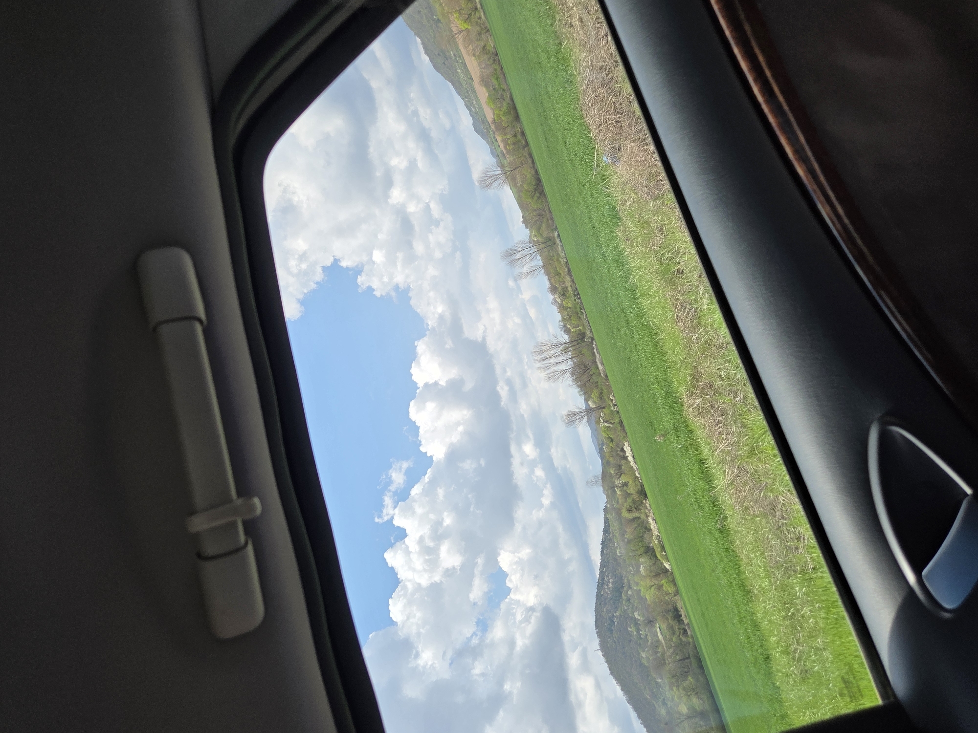 The image depicts a view from inside a vehicle, through a window. The view is of a landscape featuring a green field and a cloudy sky. A hill or low mountain is visible in the background. Parts of the car's interior are visible, including a grab handle and parts of the door or frame. The location is in Dobrodan, Bulgaria.