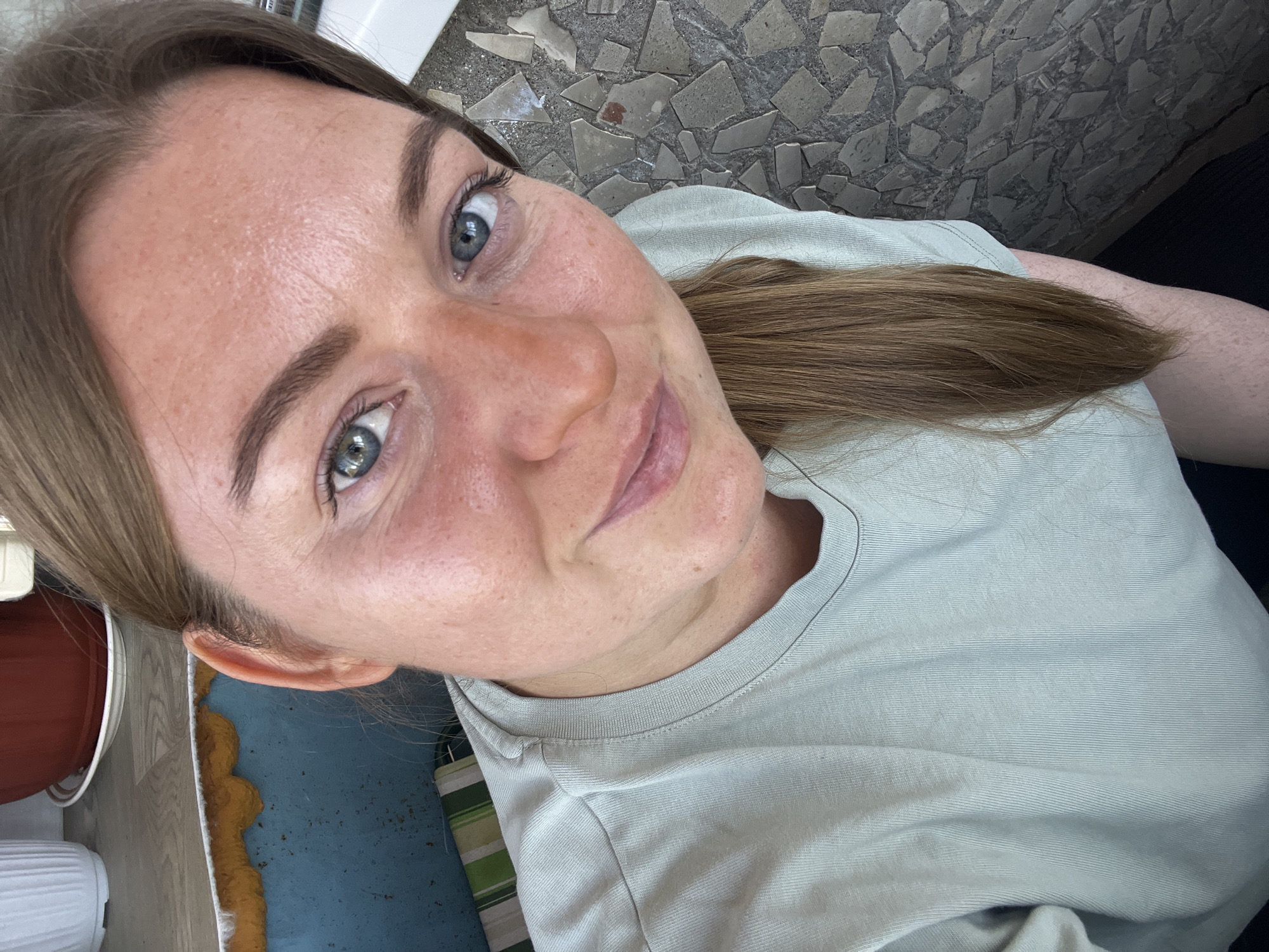 A woman with fair skin, light brown hair pulled back in a ponytail, and blue eyes is taking a selfie. She is wearing a light green t-shirt. The background includes a patterned tile floor and parts of furniture, including a blue surface with yellow and white trim. The location is Razdolye, Russia.