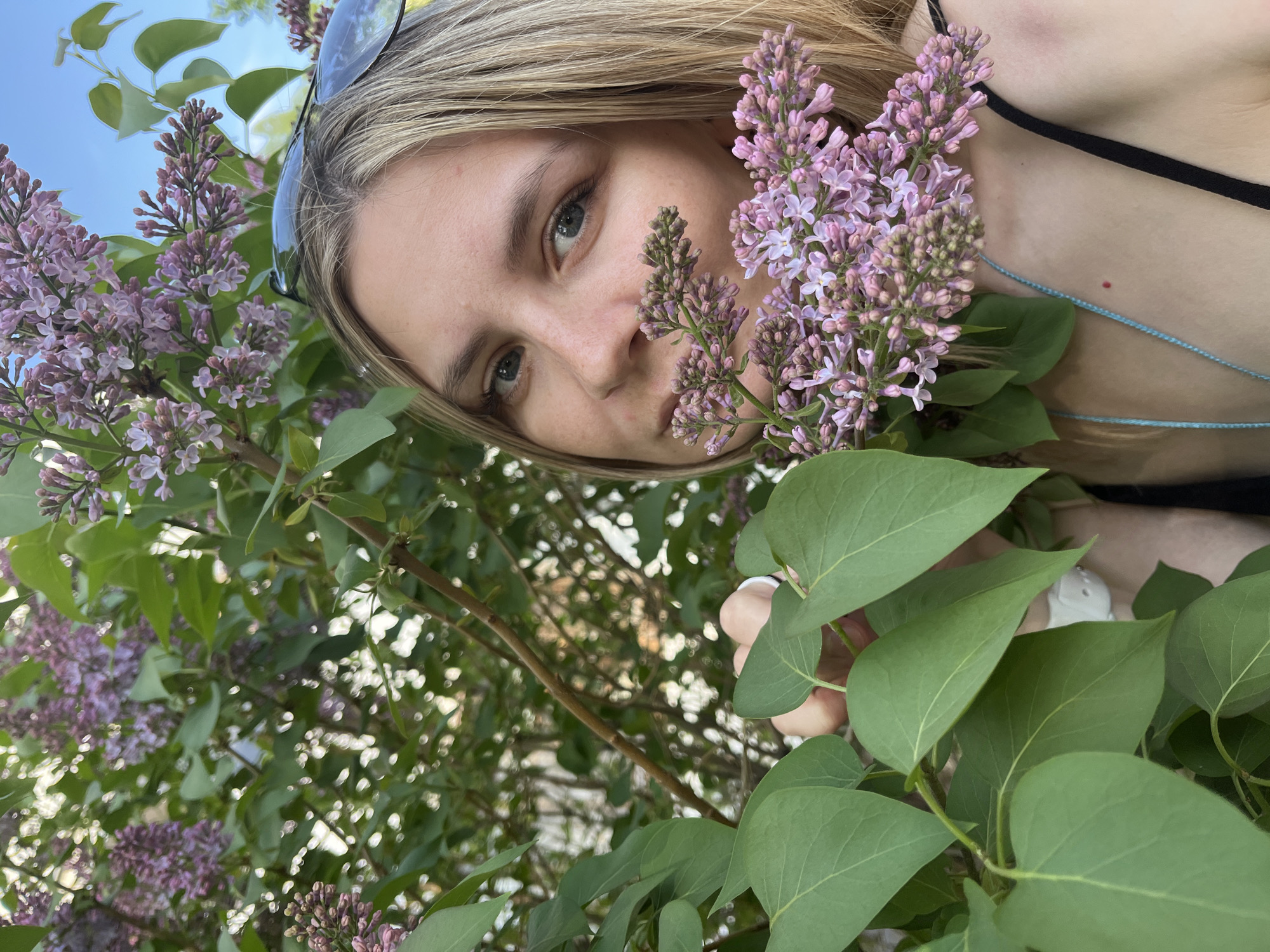 A young, blonde-haired woman with sunglasses perched atop her head poses among a lilac bush. She is wearing a black top and a thin, blue necklace. Her face is close to the lilac blossoms, which are a mix of open and closed buds. Her left hand is visible holding a leafy branch. The background is a mix of greenery and blurred structures suggesting a park or garden in the city of Chaikovsky, Russia. The sky is partly visible in the upper left corner.