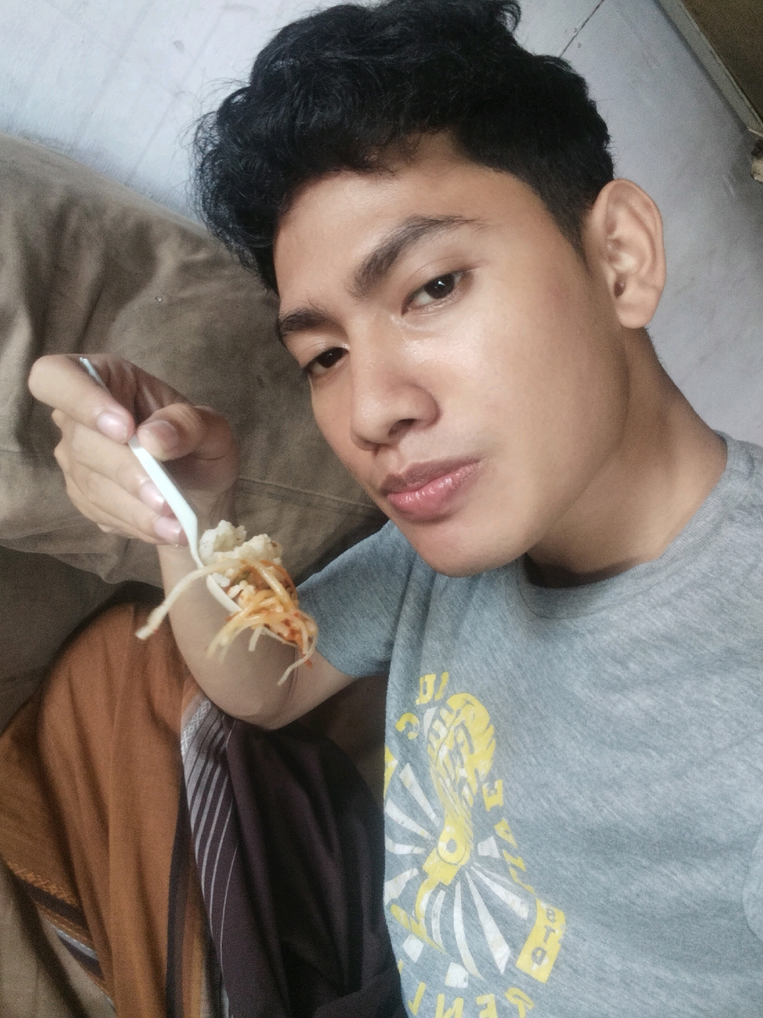 A young man with dark, curly hair is holding a white plastic fork with food on it, bringing it to his mouth. He is wearing a grey t-shirt with a yellow graphic design. The man is sitting or lying down, with a textured brown fabric in the background. The scene appears to be indoors, possibly a living room, with neutral lighting.