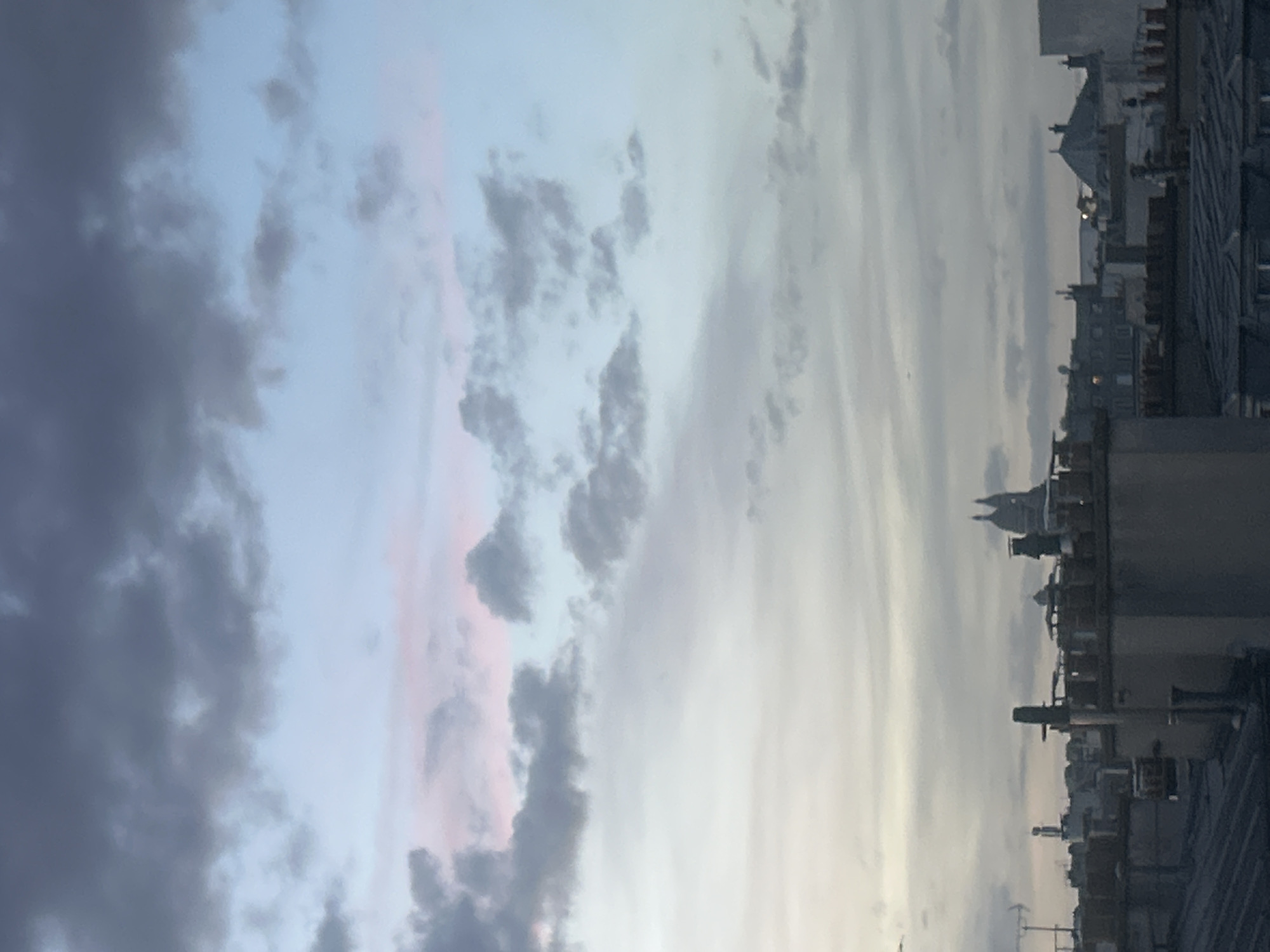 The image captures a panoramic view of a city skyline against a dramatic sky, likely at dawn or dusk. The perspective is from an elevated vantage point, looking across rooftops towards the horizon.
The sky occupies the majority of the frame, displaying a gradient of colors. On the left, dense, dark gray clouds dominate, while moving right, the sky transitions to lighter blues and soft pinks, suggesting the subtle hues of twilight or early morning. Various cloud formations, from wispy cirrus to more substantial cumulus, are scattered across the expansive sky.
Below the sky, the upper sections and rooftops of numerous buildings form a dense urban landscape. The architecture appears traditional, with pitched roofs, chimneys, and various rooftop structures typical of a European city. A highly recognizable landmark, the Sacré-Cœur Basilica, is distinctly visible towards the right side of the cityscape, characterized by its prominent white domes and spires. The buildings are silhouetted against the lighter sky, and no human activity or specific objects are clearly discernible within the city below.
The overall impression is one of tranquility and natural beauty, highlighting the blend of urban architecture with the natural spectacle of the sky at a transitional time of day. No text is visible in the image. The presence of the Sacré-Cœur Basilica strongly suggests the location is Paris, France.