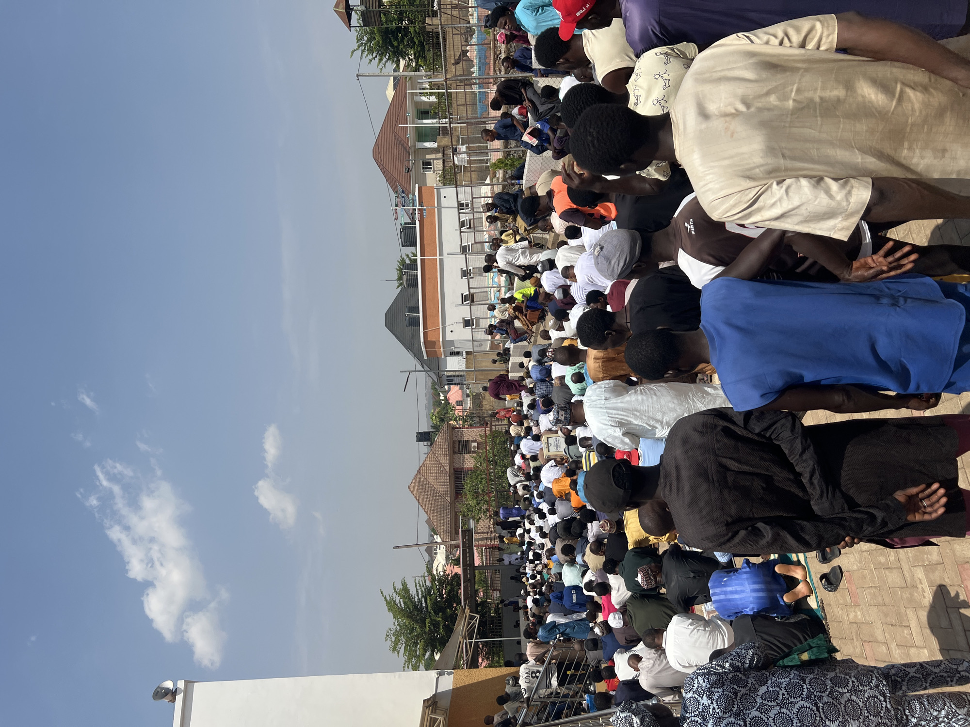 A large group of people, primarily men, are gathered outside in Abuja, Nigeria. They are kneeling and praying. Most are wearing traditional Islamic clothing. The environment includes buildings, trees, and a clear blue sky.