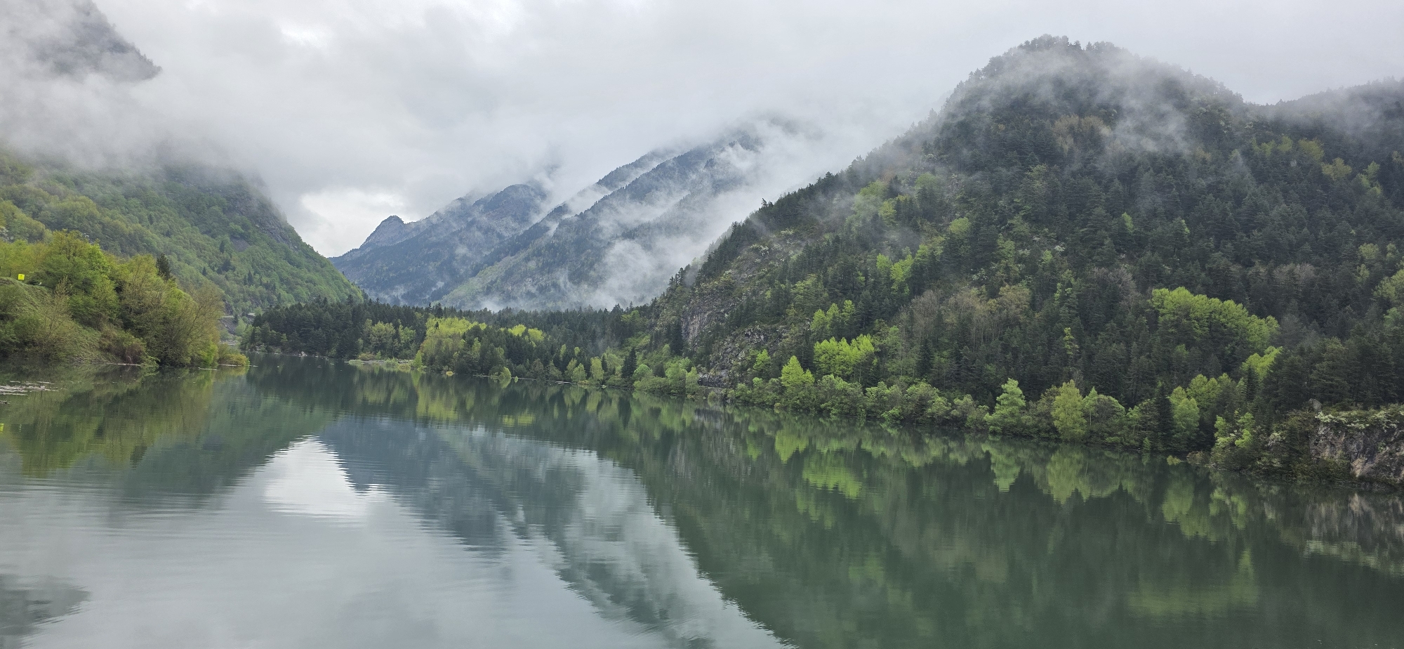 The image depicts a mountainous landscape with a lake, possibly taken in the vicinity of Benasque, Spain, though no specific landmarks are visible in the photo that confirm its exact location within the city. The foreground is dominated by the lake, which has a still surface reflecting the mountains and trees along its shores. The lake appears calm and dark in color.
The midground features the shoreline, lined with a dense collection of trees, showing a mix of dark green conifers and lighter green deciduous trees.
In the background are layers of mountains. The lower mountains have lush green vegetation, while higher peaks are shrouded in mist and clouds. The sky is overcast, contributing to the overall moody atmosphere of the scene. No people or any other sign of human activity can be observed in the image besides the possible dam on the right of the image.