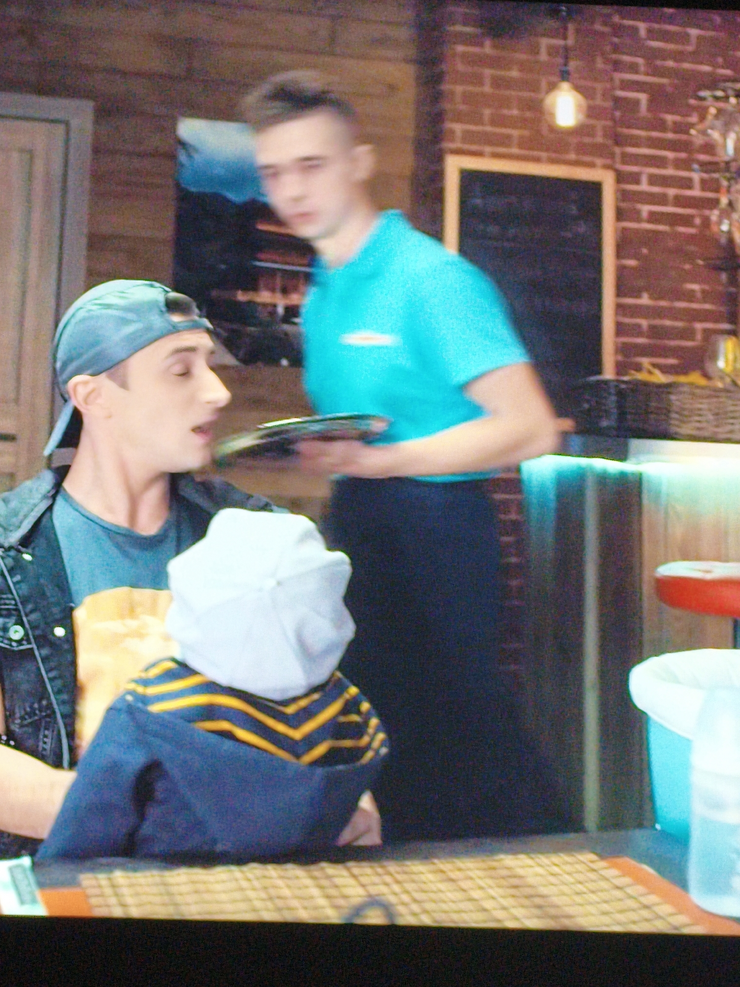 The image shows a man in a blue t-shirt and black pants standing behind a counter. He is holding a tray with a plate on it. In front of him, a man wearing a blue baseball cap and a denim jacket is looking at the tray. A child wearing a blue and yellow striped shirt and a white hat is in the foreground. The scene appears to be indoors, possibly a restaurant or cafe, with brick walls and wooden accents. There is a blackboard on the wall and a light fixture hanging from the ceiling. The lighting is dim. No specific landmarks or identifying features of Vladivostok, Russia are visible in the image.