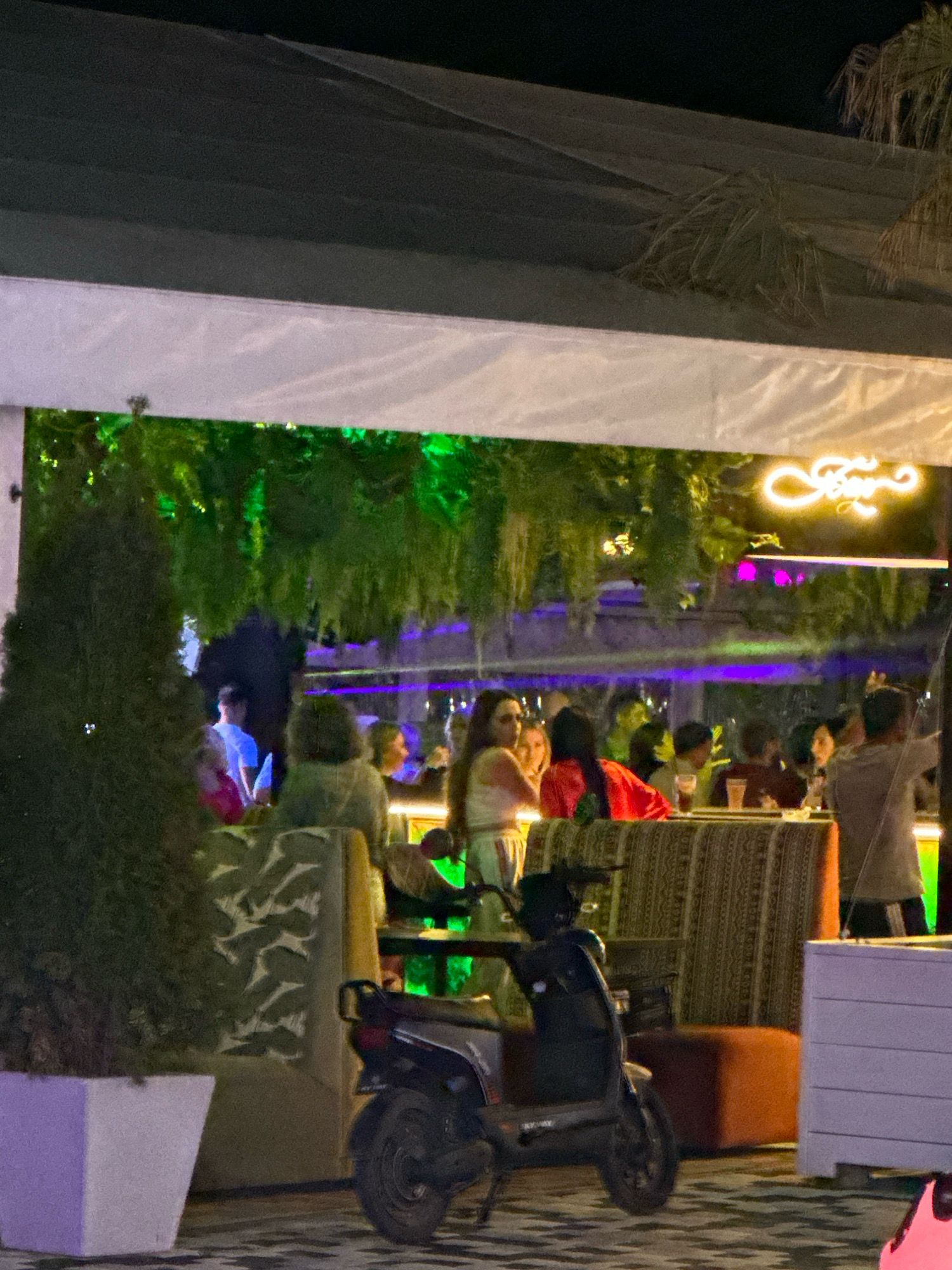 The image depicts an outdoor seating area at night, possibly a restaurant or bar, in Lermontovo, Russia. Several people are seated at tables, engaged in conversations and enjoying drinks. The ambiance is illuminated by green and purple lighting, with a neon sign visible in the background. A scooter is parked to the left of the seating area, and lush green foliage hangs from an overhead structure, contributing to the outdoor setting. The scene suggests a lively social gathering in a relaxed, nighttime environment.