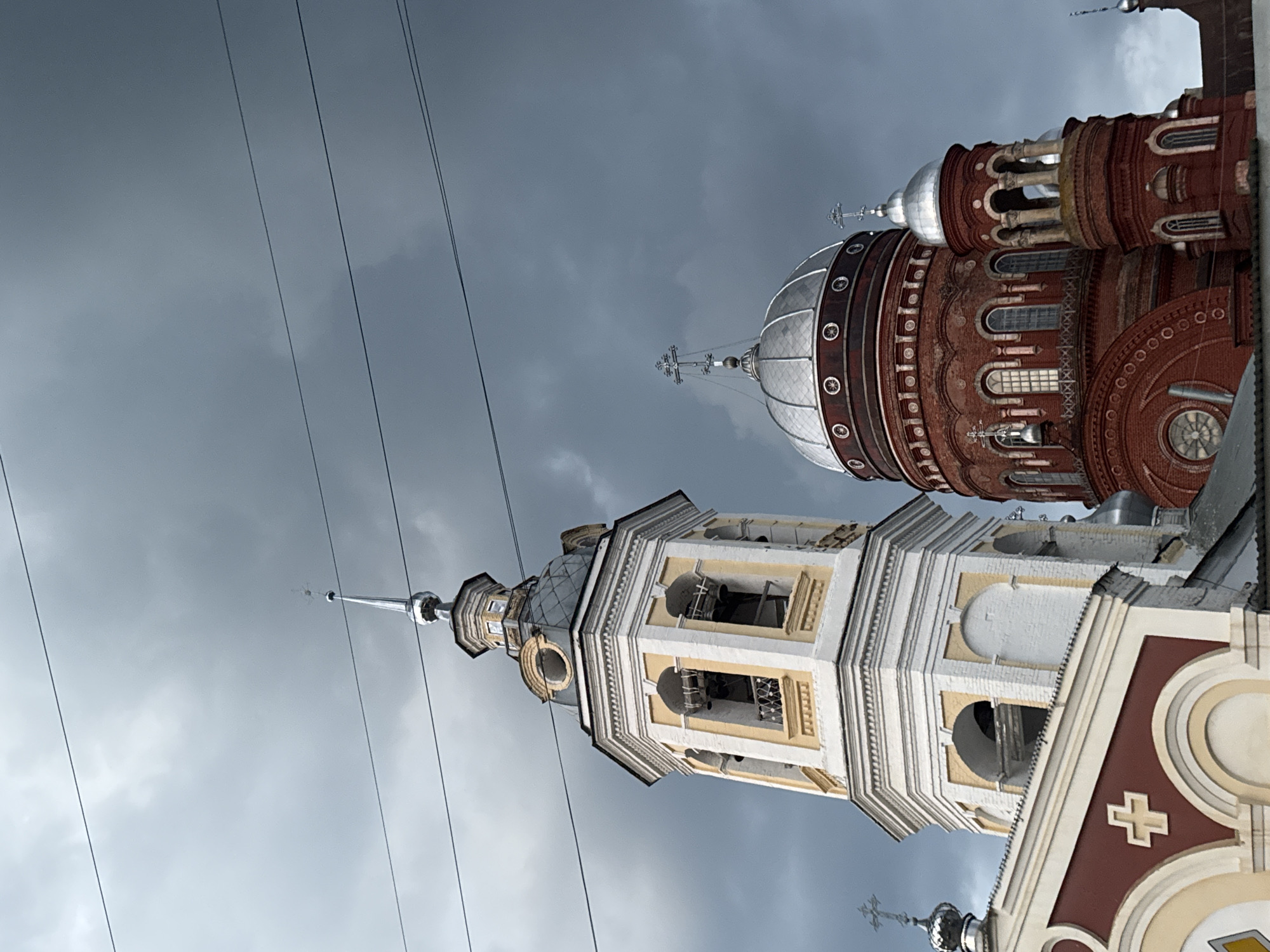 The image depicts the bell tower and domes of the Holy Trinity Cathedral in Urzhum, Russia, under a cloudy sky. The bell tower is tall and octagonal, painted white and yellow, with arched openings revealing bells. Beside it, a large, red brick dome with a silver, onion-shaped roof stands prominently, adorned with decorative elements and topped with a cross. Smaller red brick structures with silver domes are also visible. The scene is framed by overhead power lines, adding a modern contrast to the historic architecture. The overall atmosphere is somber due to the overcast sky.