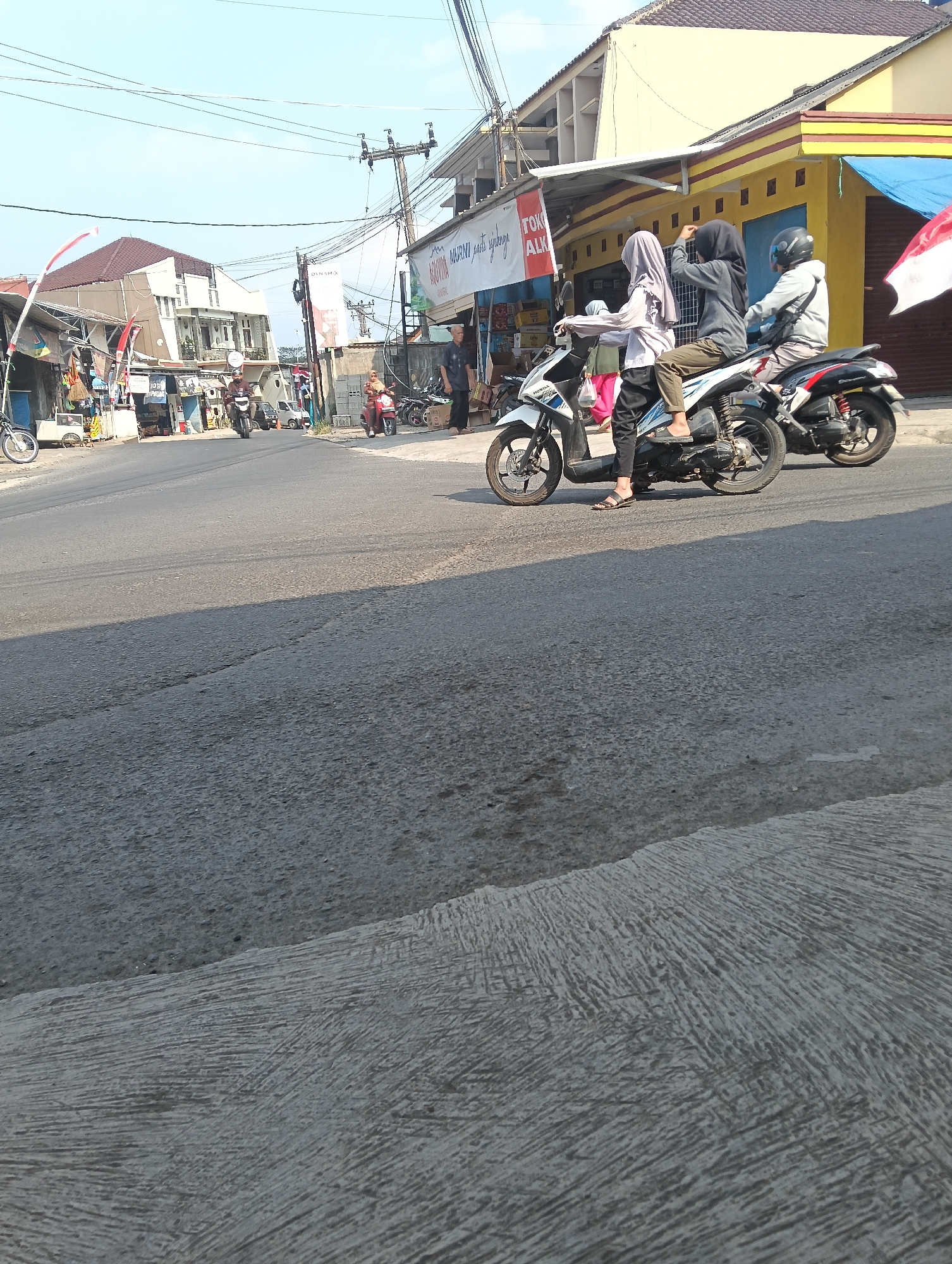 The image shows a street scene in Puspahiang, Indonesia, with several motorcycles and people. In the foreground, the asphalt road shows wear and tear, with distinct lines and patches. On the right side of the road, there are buildings with yellow and red facades. A group of people are on two motorcycles. The first motorcycle, a white Honda, carries three individuals. The rider is wearing a grey long-sleeved shirt and dark pants, with a black helmet. The middle passenger, a female, is wearing a grey hijab and a grey long-sleeved top with dark pants. The person behind her, also a female, wears a light purple hijab and a matching top. The second motorcycle, a dark-colored scooter, is positioned behind the first, with its rider wearing a grey hoodie and a black helmet.
In the background, on the left side of the street, there are more buildings and shops. One building has a prominent brown roof and white and beige walls. Various stalls and shops line the street, with some displaying merchandise. A red and white Indonesian flag is visible on a pole in front of one of the buildings. Several other motorcycles are parked or moving along the street. Power lines crisscross the sky. The lighting suggests it is daytime, with the sun casting shadows. The overall environment appears to be a typical urban or semi-urban street in Indonesia.
