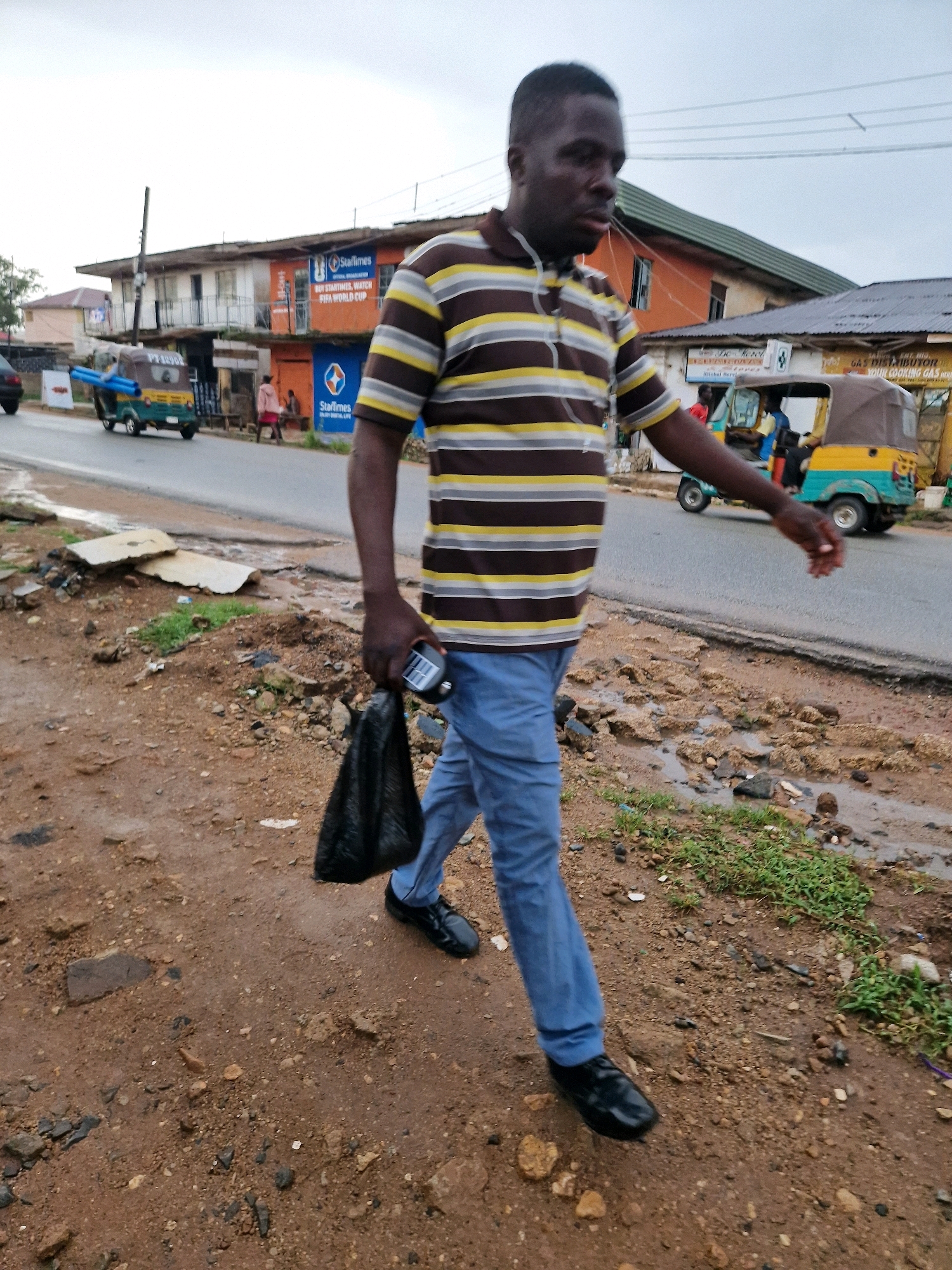 A man, wearing a brown, yellow, and white striped polo shirt and blue trousers, walks down a dirt path. He is carrying a black plastic bag in his right hand and a black object, possibly a mobile phone, in his left hand. The ground is a mixture of dirt, gravel, and some green grass. In the background, a paved road is visible, with several vehicles, including a tuk-tuk and another three-wheeled vehicle. Buildings line the road, some of which appear to be shops. The sky is overcast. The scene is likely in Jos, Nigeria, based on the visual cues such as the architecture, vehicles, and the general environment.