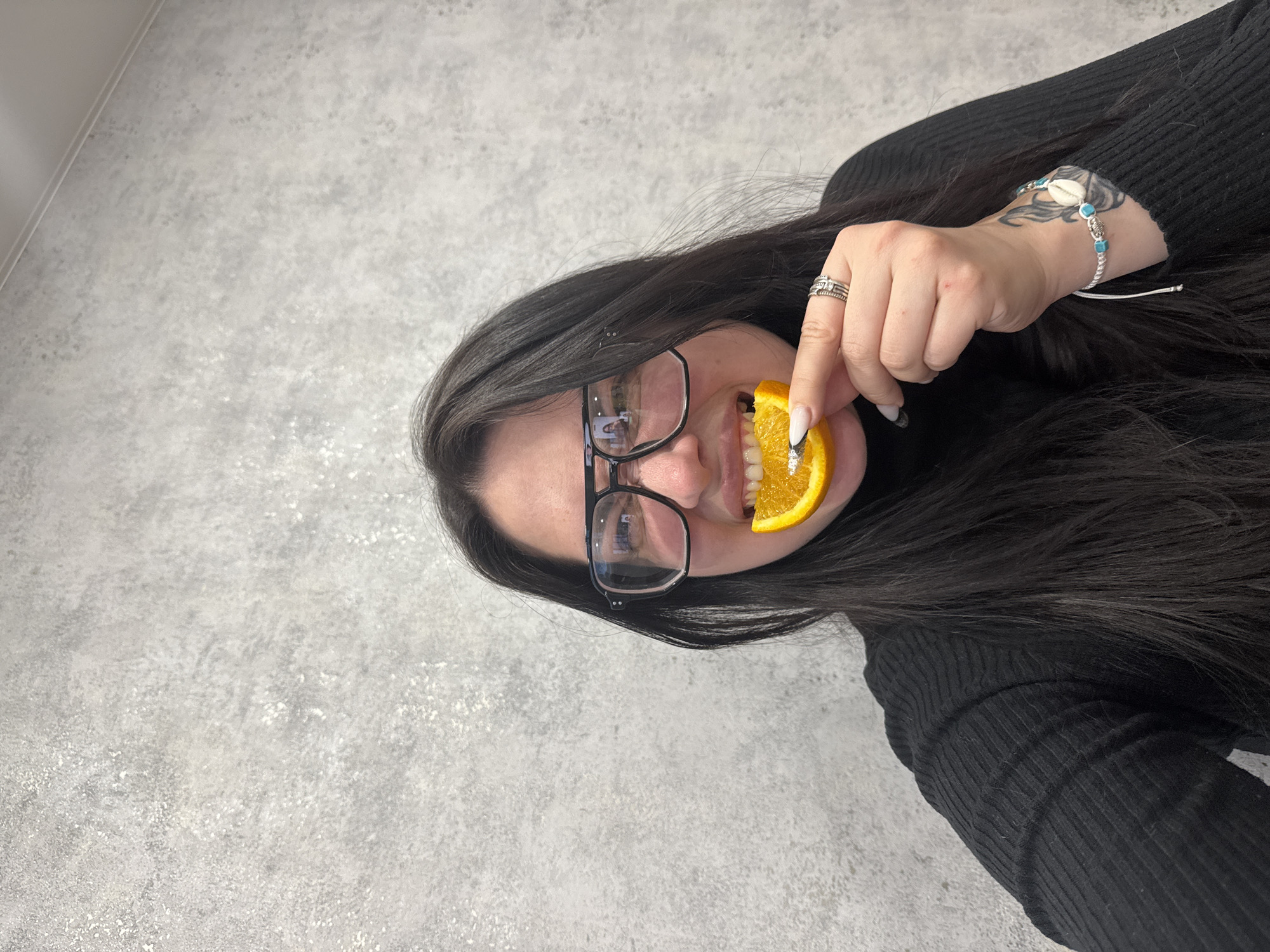 A medium close-up shot captures a woman holding a slice of orange playfully to her mouth. She has long, dark hair that cascades around her shoulders and is wearing a black, ribbed long-sleeved top. Her face is partially visible behind the orange slice, and she is wearing dark-framed glasses. A faint reflection, possibly of a screen or another person, can be seen in the lenses of her glasses, though no discernible text is visible.
On her left hand, she wears several rings on her index and ring fingers, and her nails are manicured with white tips and a glitter accent on one nail. A bracelet featuring a cowrie shell and small turquoise beads is worn on her left wrist, above which a tattoo is visible on her forearm. The woman appears to be smiling or making a playful expression as she holds the fruit.
The background is a textured light gray wall with subtle patterns and variations, suggesting an indoor setting. A faint line in the upper left corner indicates a wall or ceiling junction. The lighting is even and bright, highlighting the subject. The overall impression is casual and cheerful.
