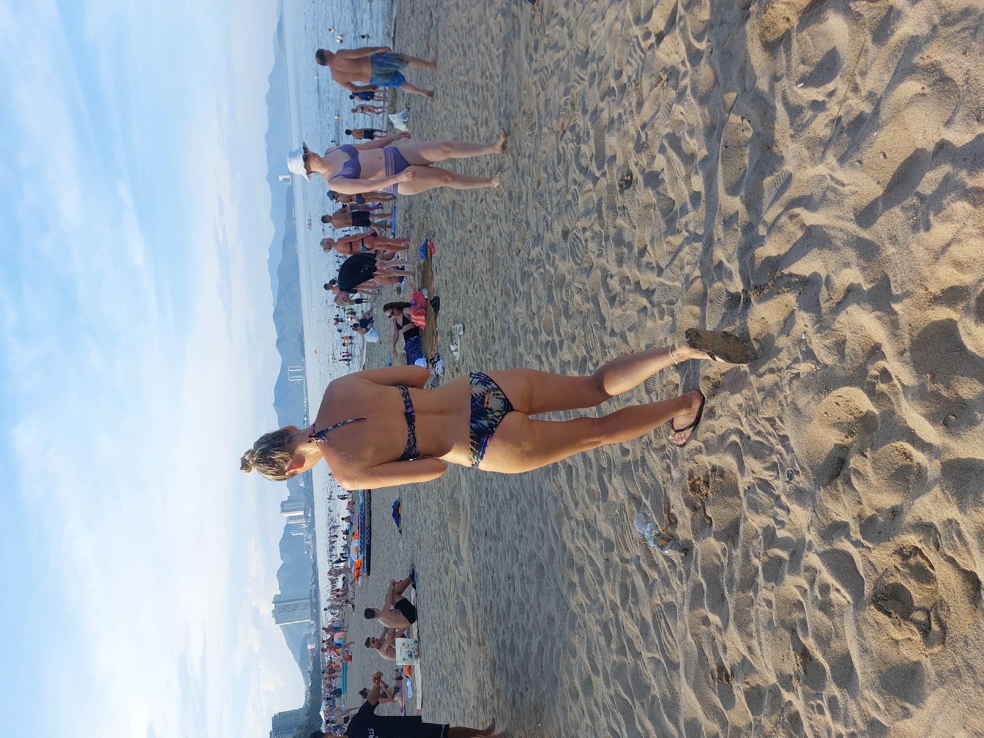 The image shows a busy beach scene in Nha Trang, Vietnam. The sand is a light tan color, with various footprints and ripples from the wind. In the foreground, a woman with her back to the camera is walking on the sand. She has blonde hair pulled up in a bun and is wearing a patterned bikini. Her feet are bare except for a dark thong sandal on her left foot. To her right, a plastic water bottle and some trash are visible on the sand.
In the middle ground, a large number of people are spread across the beach and in the shallow water. Several individuals are wearing swimsuits, and some are sitting or lying on towels. There are several inflatable rafts visible. A woman in the middle ground on the right is wearing a purple bikini and a white bucket hat. Further back, a man in blue swim trunks is walking away from the camera. The ocean is a calm blue, with many people swimming and wading in the water.
The background features a wide expanse of the ocean with a distant shoreline dotted with tall buildings and mountains. The sky is a pale blue with scattered clouds. The overall impression is a lively summer day at a popular tourist destination.
