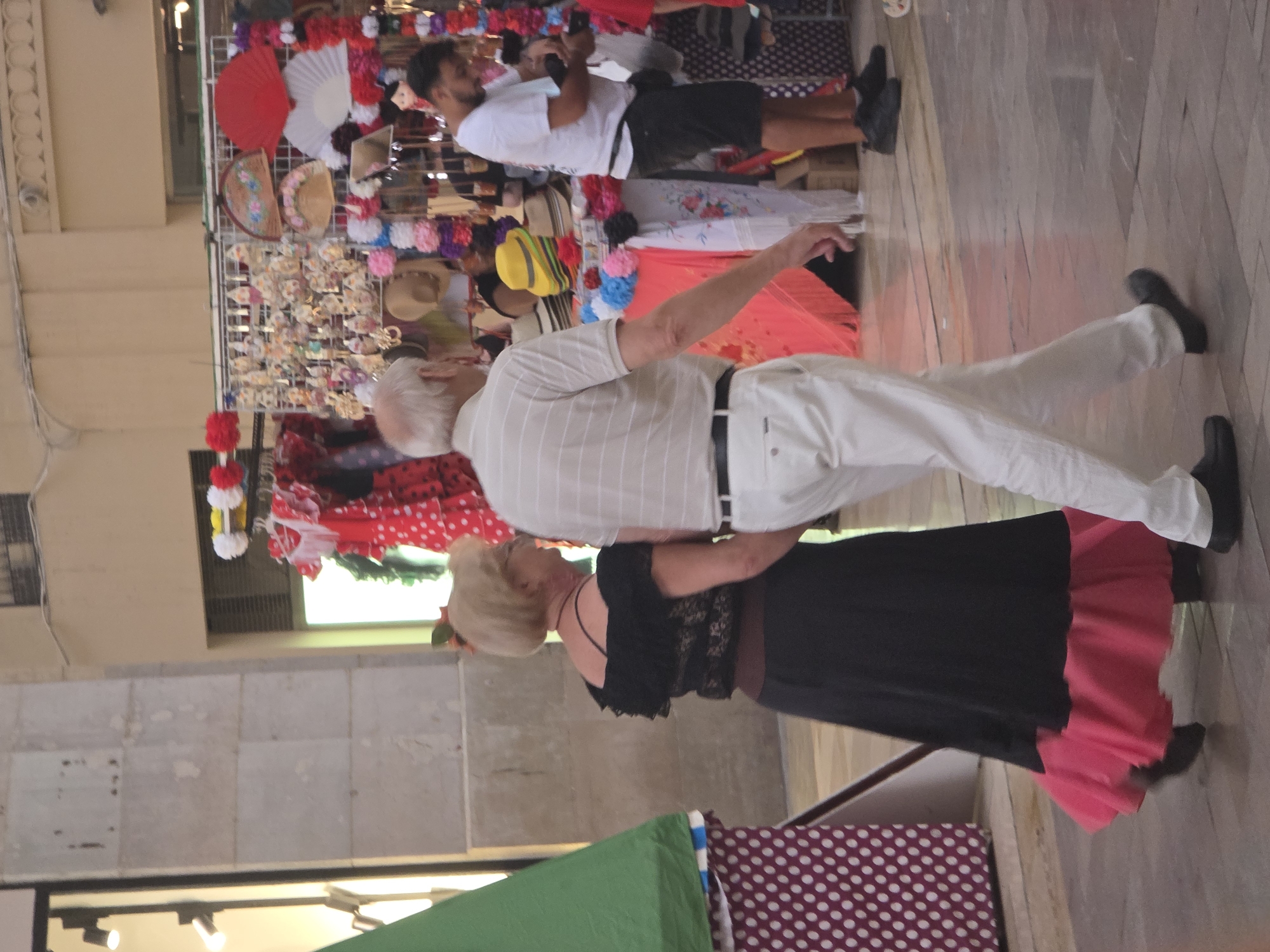 The image depicts a street scene in Malaga, Spain, likely on a warm day, as indicated by the attire of the people and the open stalls. In the foreground, a couple, an older white man and an older white woman, are walking away from the viewer. The man is wearing a light-colored, horizontally striped t-shirt and white trousers. The woman is wearing a black off-the-shoulder top with lace detail and a long black skirt with a bright pink ruffled hem. To the right, a young man with dark hair and a beard is visible, wearing a white t-shirt and dark shorts, holding a dark object in his hands. Behind the main subjects, there is a market stall displaying various items, including colorful fans, hats, and what appear to be decorative ornaments, some of which are red and white. A green tent-like structure is partially visible on the left side of the frame, with a polka-dot pattern on its base. The ground is paved with stone tiles. The architecture in the background suggests a Spanish urban environment. The lighting suggests it is daytime, possibly late afternoon or early evening, with soft, diffused light.