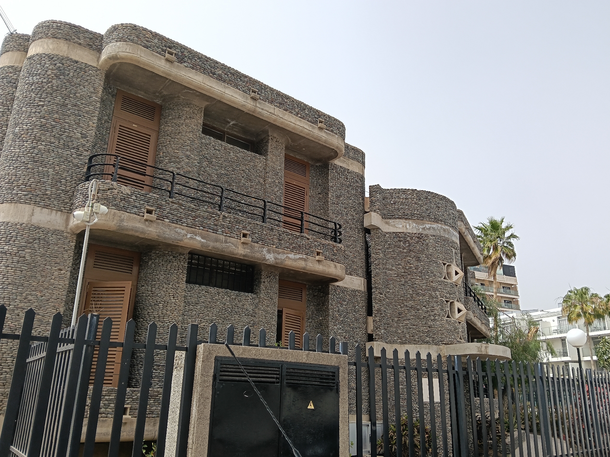 A low-angle, full-color shot depicts a building in San Bartolomé de Tirajana, Spain, characterized by an exterior constructed from small, rounded stones set in concrete. The structure is behind a black, wrought-iron fence with vertical bars, a low concrete wall, and a utility box with black metal doors, featuring a yellow electrical hazard symbol.
The building has a multi-level design with rounded corners. Shutters cover the windows. The building features a balcony with a black iron railing on the second floor. A palm tree is positioned to the right of the building. The sky is overcast. In the background, parts of other buildings can be seen.