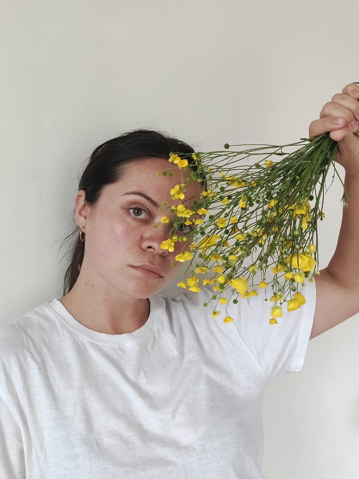 A medium shot captures a woman holding a large bunch of yellow wildflowers, possibly buttercups, in front of her face, partially obscuring her features. Her dark hair is pulled back, revealing a simple gold hoop earring in her left ear. Her left eye is visible, looking directly at the viewer with a neutral, perhaps thoughtful, expression. Her skin appears natural with a healthy glow. She is wearing a plain white short-sleeved t-shirt with a round neckline. The flowers, with their vibrant yellow petals and green stems, dominate the right side of the frame. The background is a simple, light off-white wall, suggesting an indoor setting with soft, even lighting. There is no visible text.