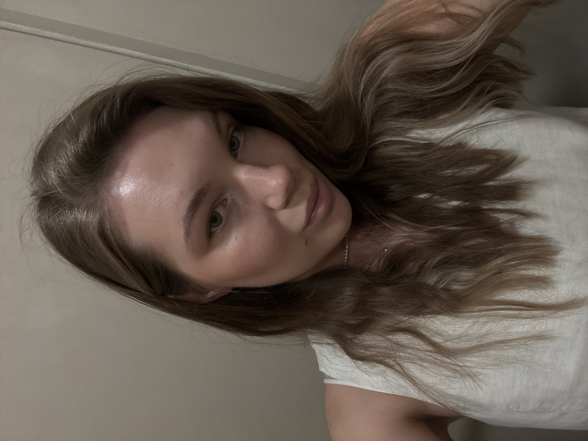 This is a selfie of a young woman with long, light brown hair, taken indoors against a neutral-colored wall. She is wearing a white sleeveless top and a delicate necklace. Her eyes are green, and she is looking directly at the camera with a slight smile. The lighting suggests an indoor environment, possibly a room in a residential building in the Vračar Urban Municipality, Serbia, given the context. There are no other people or objects of note in the frame, and the focus is entirely on the woman's face and upper body.