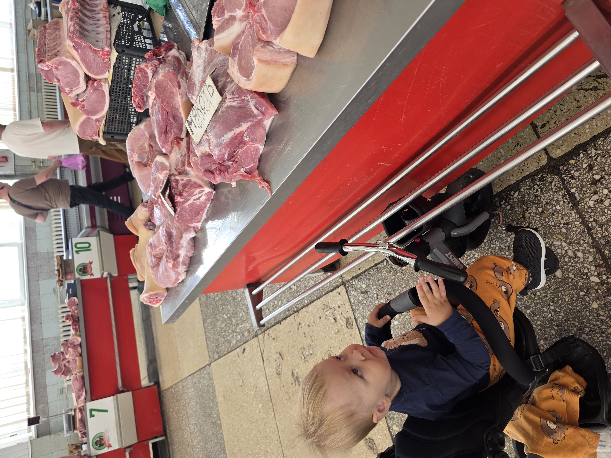 The image depicts a market scene where various cuts of raw meat, primarily pork, are displayed on a metal counter. A child is visible in a stroller, looking towards the left of the frame. The child is wearing a navy blue long-sleeved shirt and orange pants with a bear print. The stroller has a black frame and a black seat.
In the background, several market stalls are set up, with more meat displayed. Some stalls have numerical labels, including "10" and "1" with accompanying graphics. The environment appears to be an indoor market or a covered outdoor market with tiled flooring. The lighting suggests it is daytime.
There is a sign visible on some of the meat that appears to be in Cyrillic script. One sign, in particular, has the word "МЯСО" (meat) written on it.
The overall scene conveys a typical market environment where food products are being sold. The presence of the child in the stroller suggests a family outing or shopping trip.