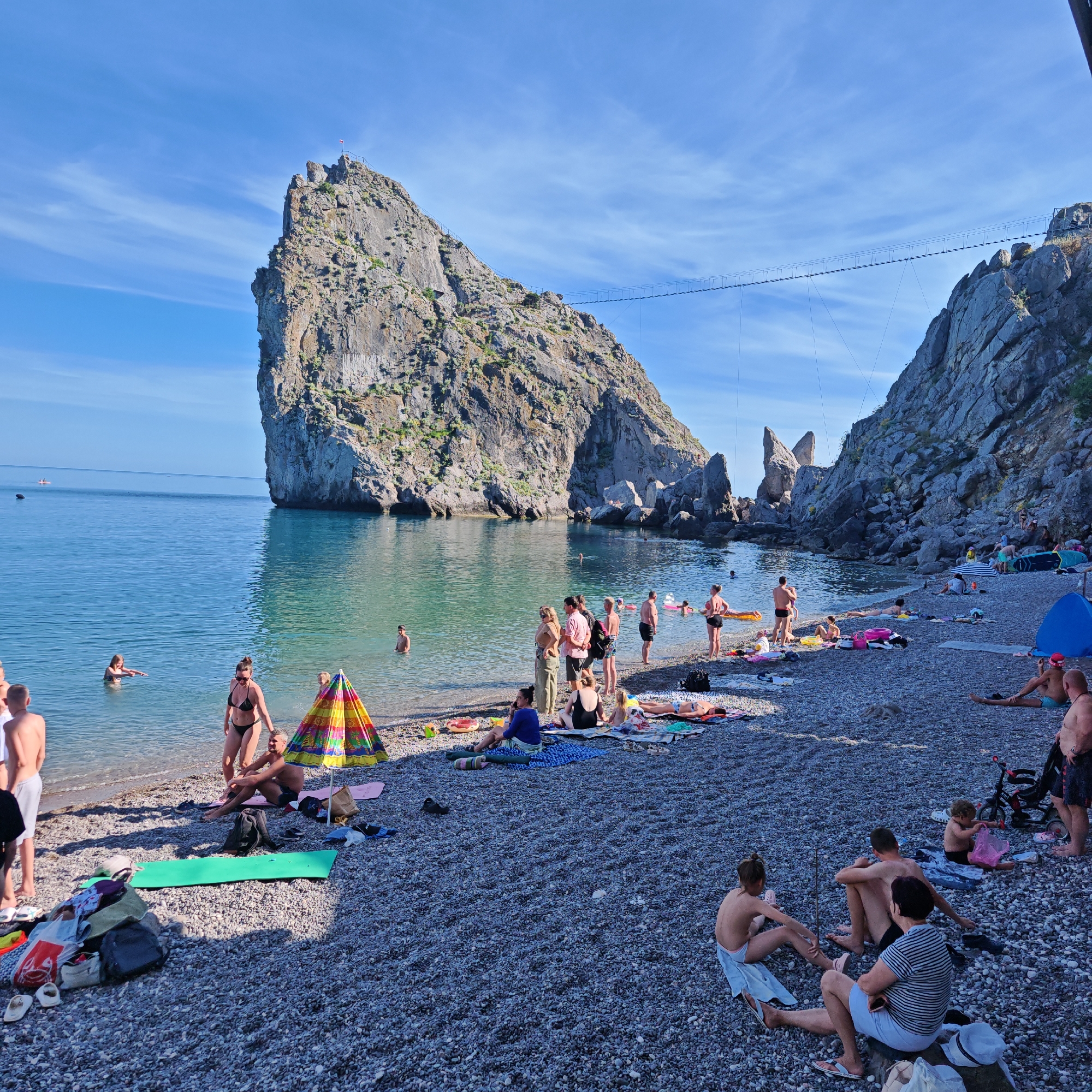 This is a wide-angle shot of a crowded pebble beach on a sunny day, with a large, rugged rock formation jutting out into the clear, blue sea. The time of day appears to be late morning or early afternoon, given the bright sunlight and clear blue sky with wispy clouds. The water is calm and inviting, with several people swimming and wading near the shore.
The beach is populated with numerous individuals and groups relaxing, sunbathing, and socializing. People are dressed in swimwear, and various beach accessories like towels, umbrellas, inflatable toys, and bags are scattered across the pebbled shore. A colorful umbrella stands out near the center of the frame. Some individuals are seated on towels, while others are standing or walking. A suspension bridge is visible spanning between two rocky outcrops in the background, suggesting a dramatic landscape. The overall atmosphere is one of leisure and enjoyment on a summer day at the coast. The location appears to be a popular tourist spot, likely in a rocky coastal area.