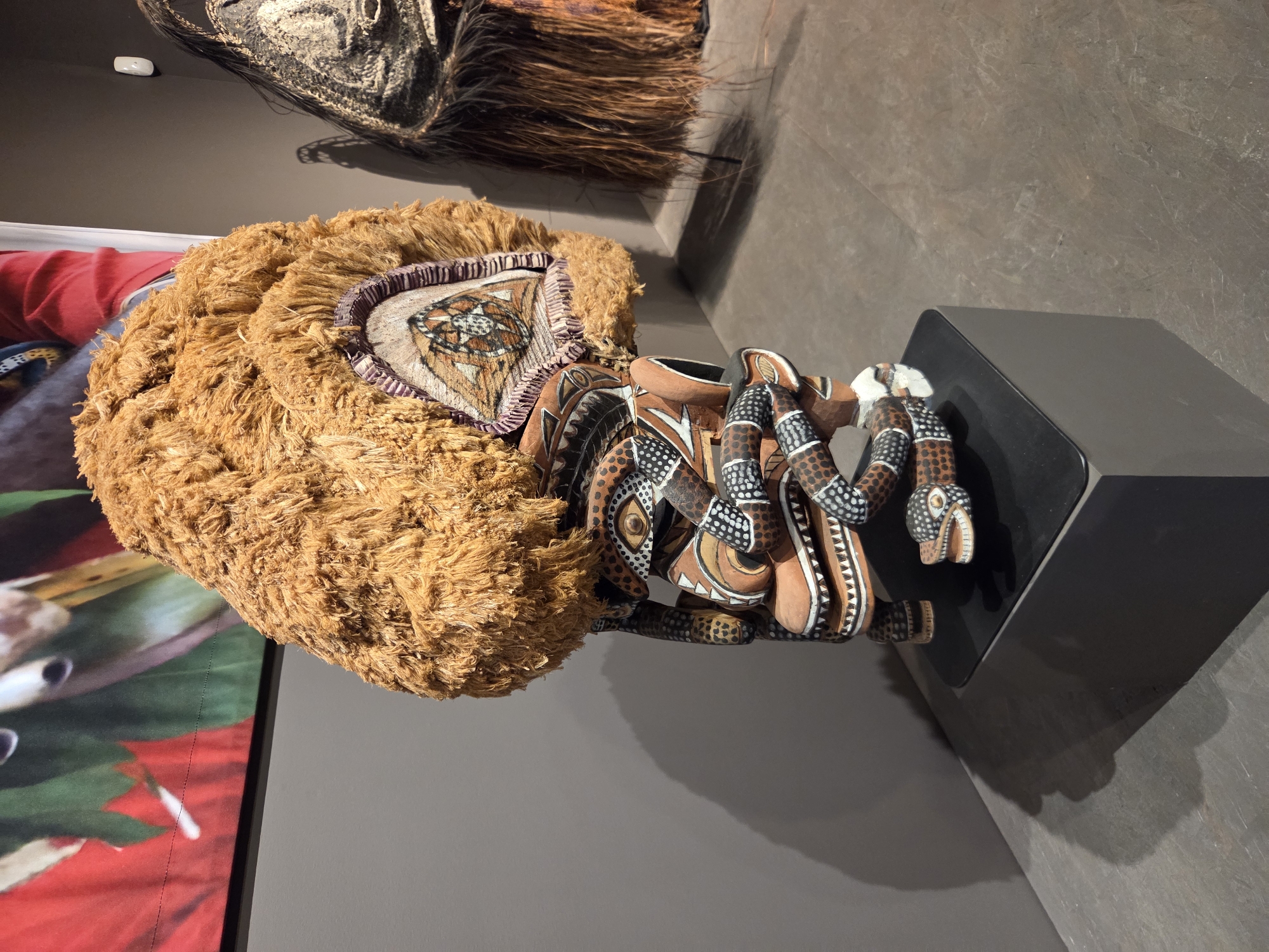 The image shows a close-up view of an elaborately carved wooden mask on display in an exhibition. The mask features a large, furry headdress, intricate facial details with painted eyes, and snake-like elements coiled around the face. The mask is mounted on a black pedestal, which is itself placed on a larger gray cube pedestal. The background consists of a gray wall and a portion of a large, colorful fabric art piece hanging on the wall. To the right of the mask, another exhibit with dark, fringed material is partially visible. The lighting casts shadows on the wall and floor, suggesting an indoor museum or gallery setting. This artwork is located at the Bowers Museum in Santa Ana, California, United States of America.